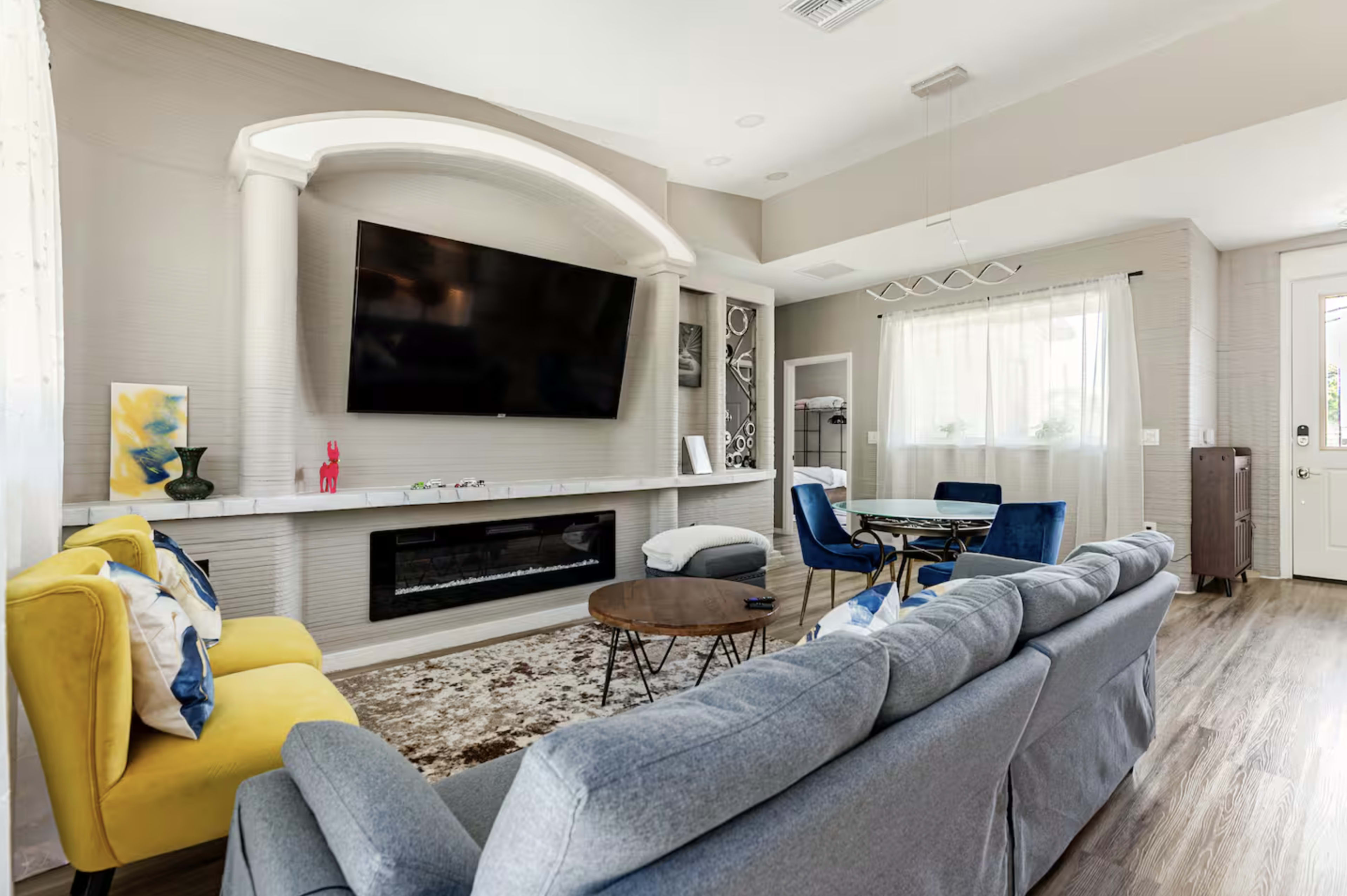 The image shows a modern living room featuring a gray couch, yellow accent chair, a wall-mounted television, and a dining area with a round table.