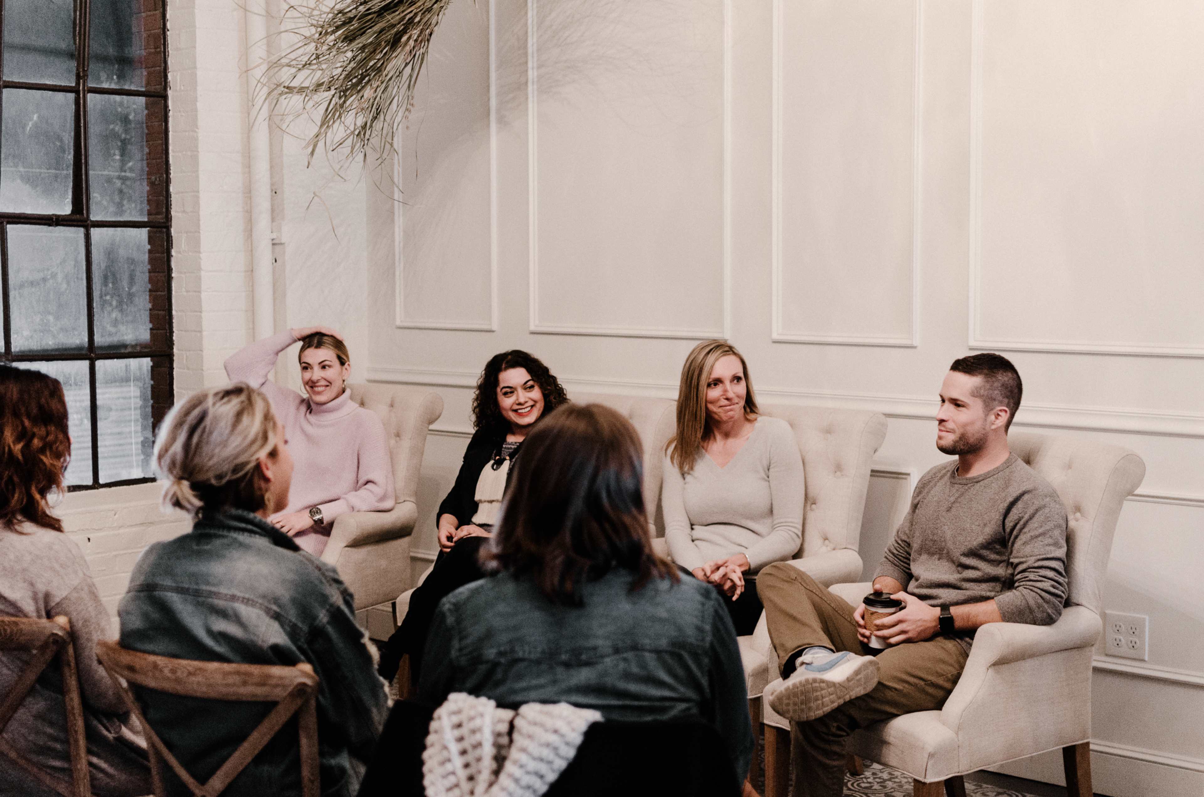 A group of six people sits in a cozy, well-lit room with large windows and soft furnishings, engaged in conversation.
