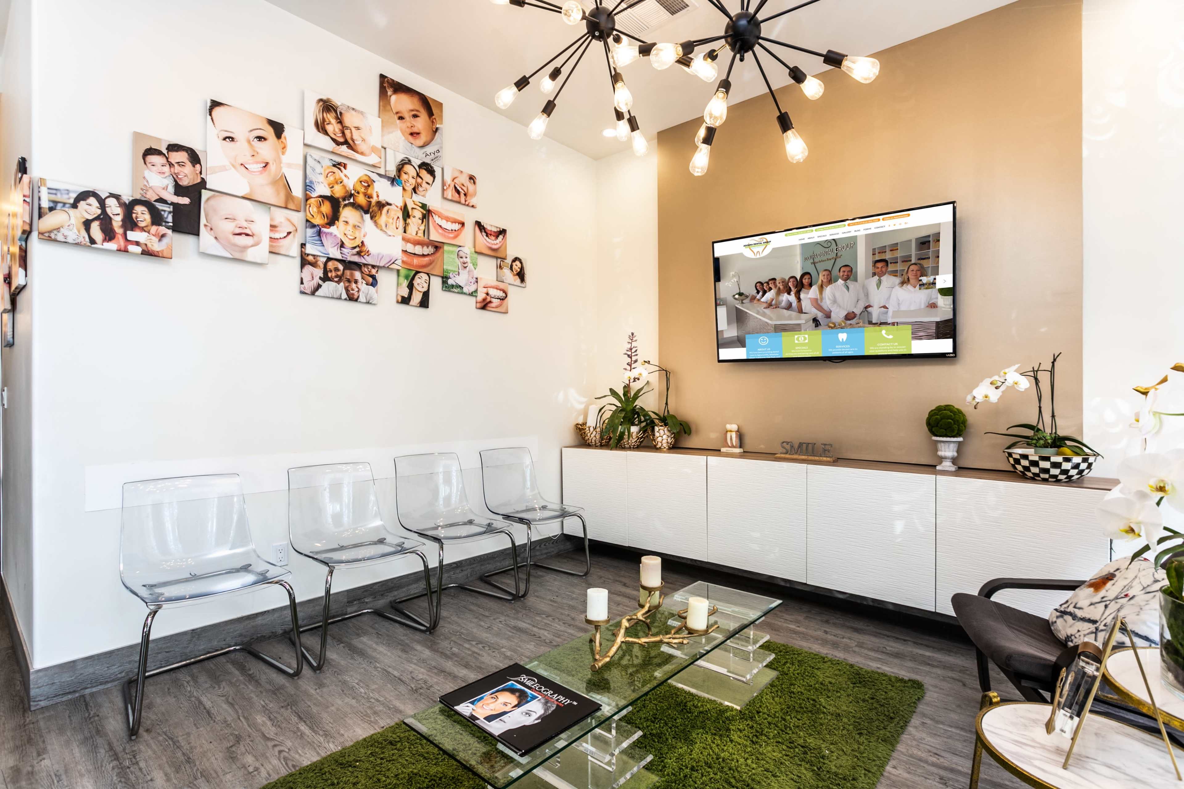 A waiting room with transparent chairs, a wall decorated with family photos, and a large screen displaying information, set against a backdrop of light-colored walls and a green rug.