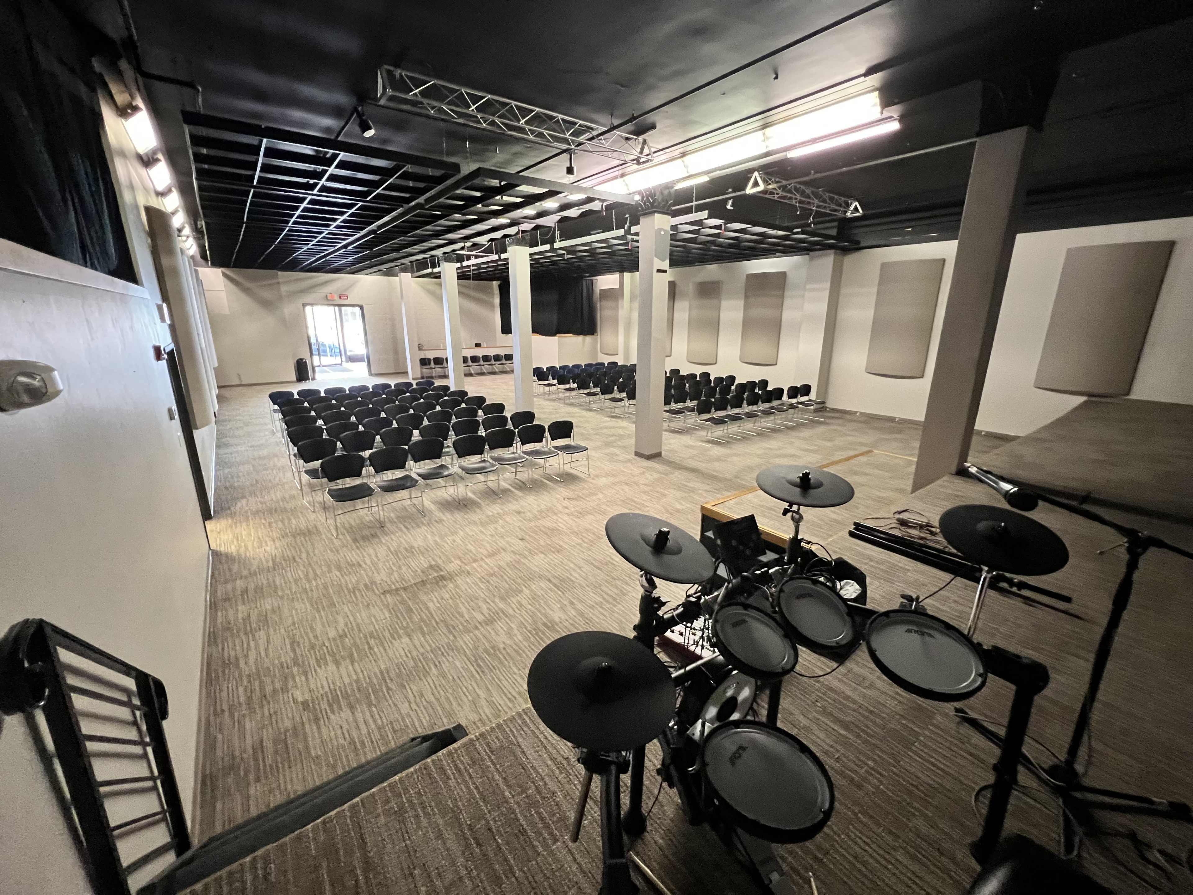 A spacious auditorium with rows of black chairs facing a stage area equipped with an electronic drum set.