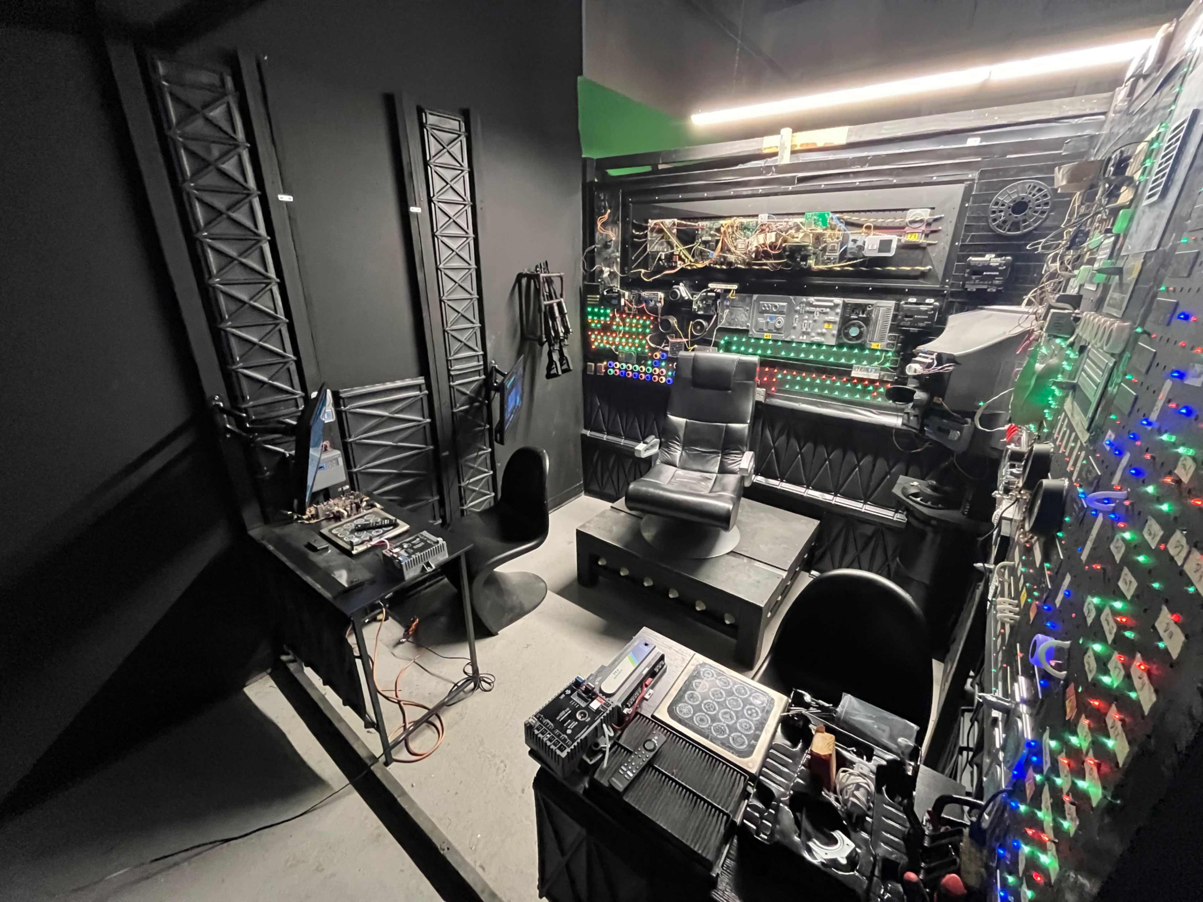 The image shows a dimly lit control room with a high-backed chair, various electronic equipment, and a wall covered in colorful buttons and lights.