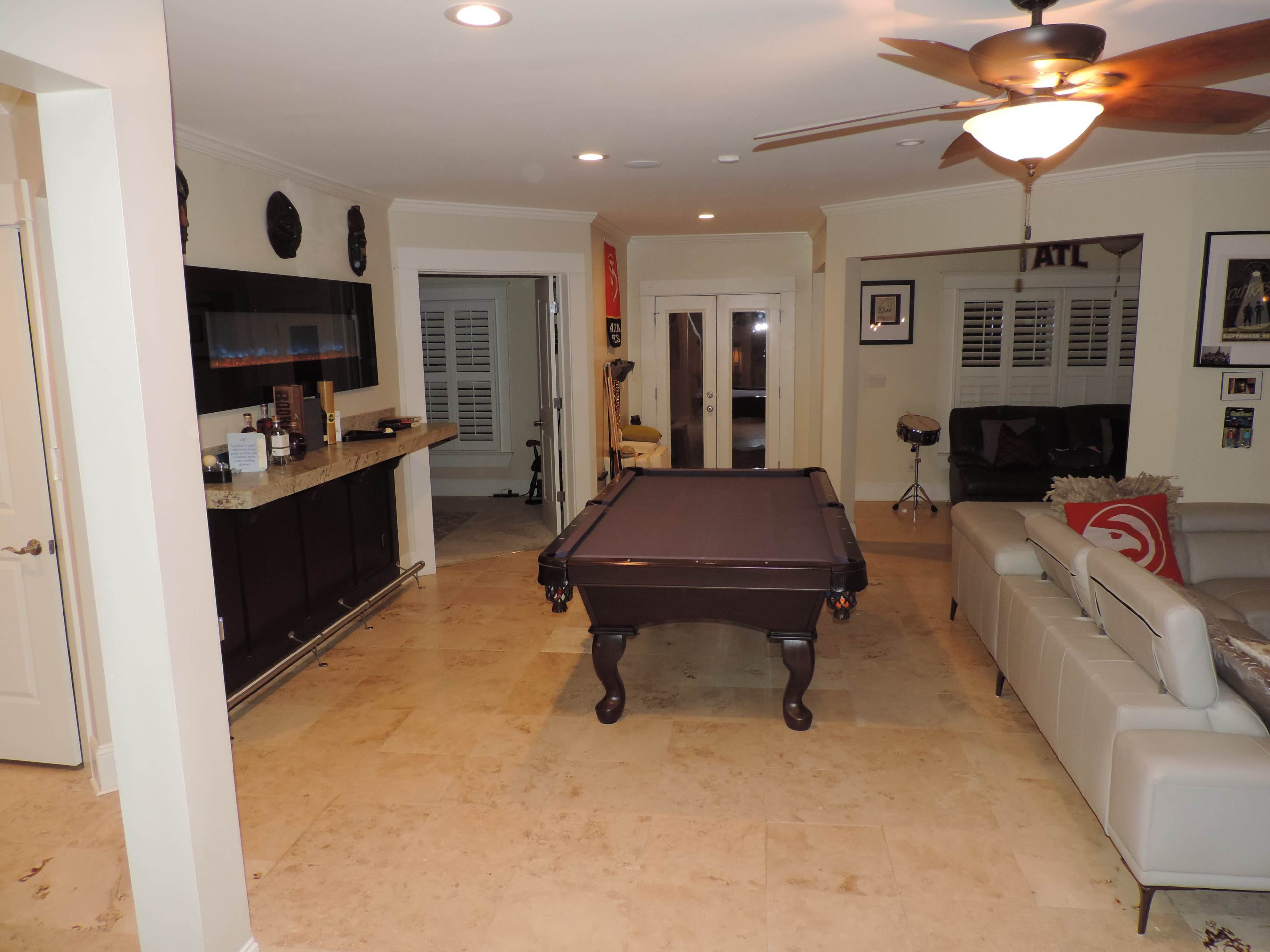 The image shows a spacious room featuring a billiard table at the center, a bar area with a countertop on the left, and a seating area with a light-colored sofa on the right.