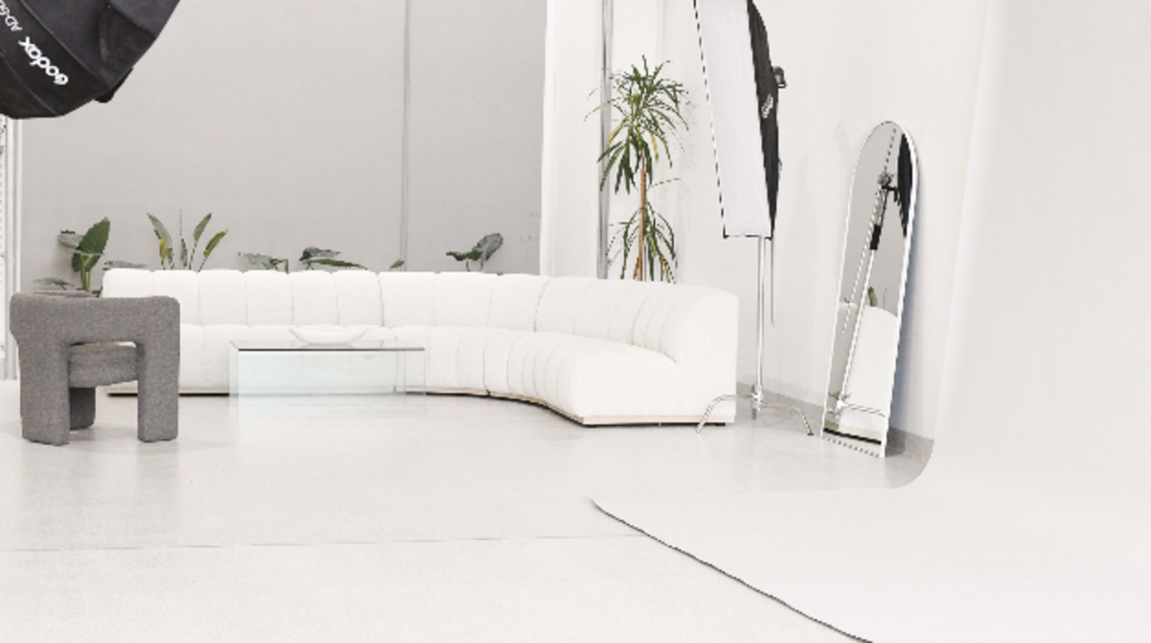 A minimalist studio space with a white curved sofa, a glass coffee table, a full-length mirror, and a backdrop, accompanied by potted plants.