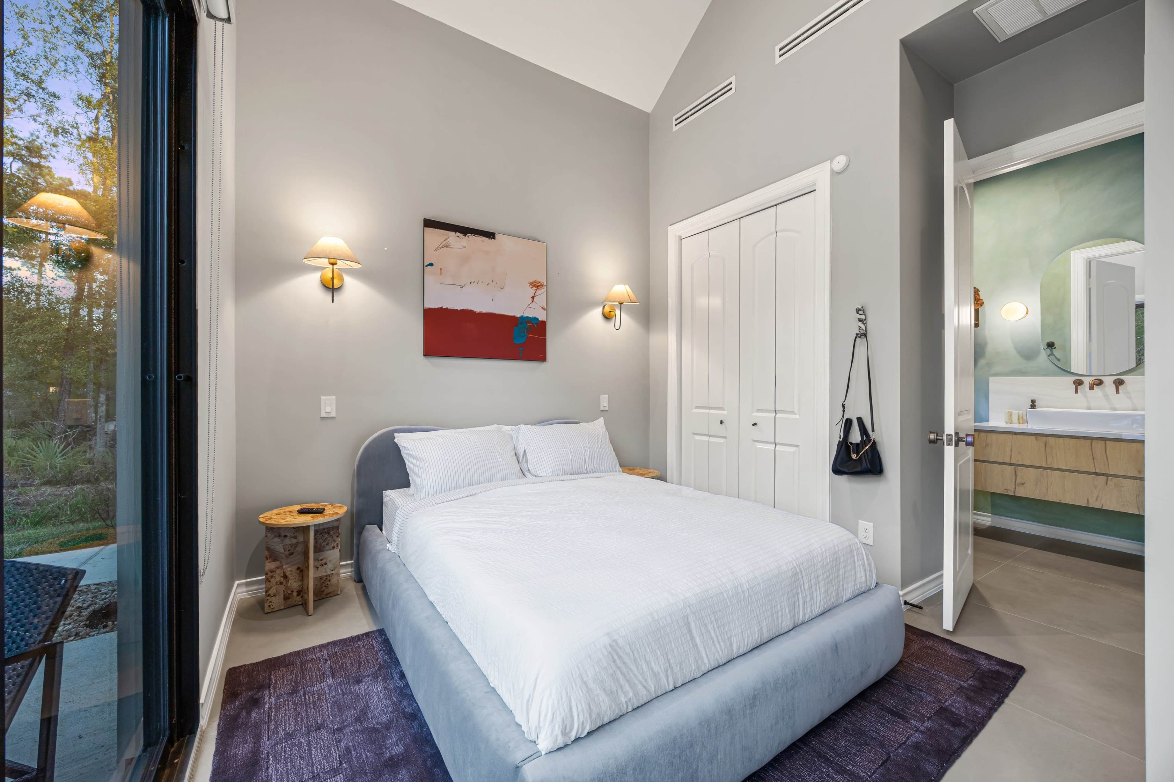 The image shows a modern bedroom featuring a bed with white linens, a wall-mounted artwork, and a doorway leading to a bathroom area.