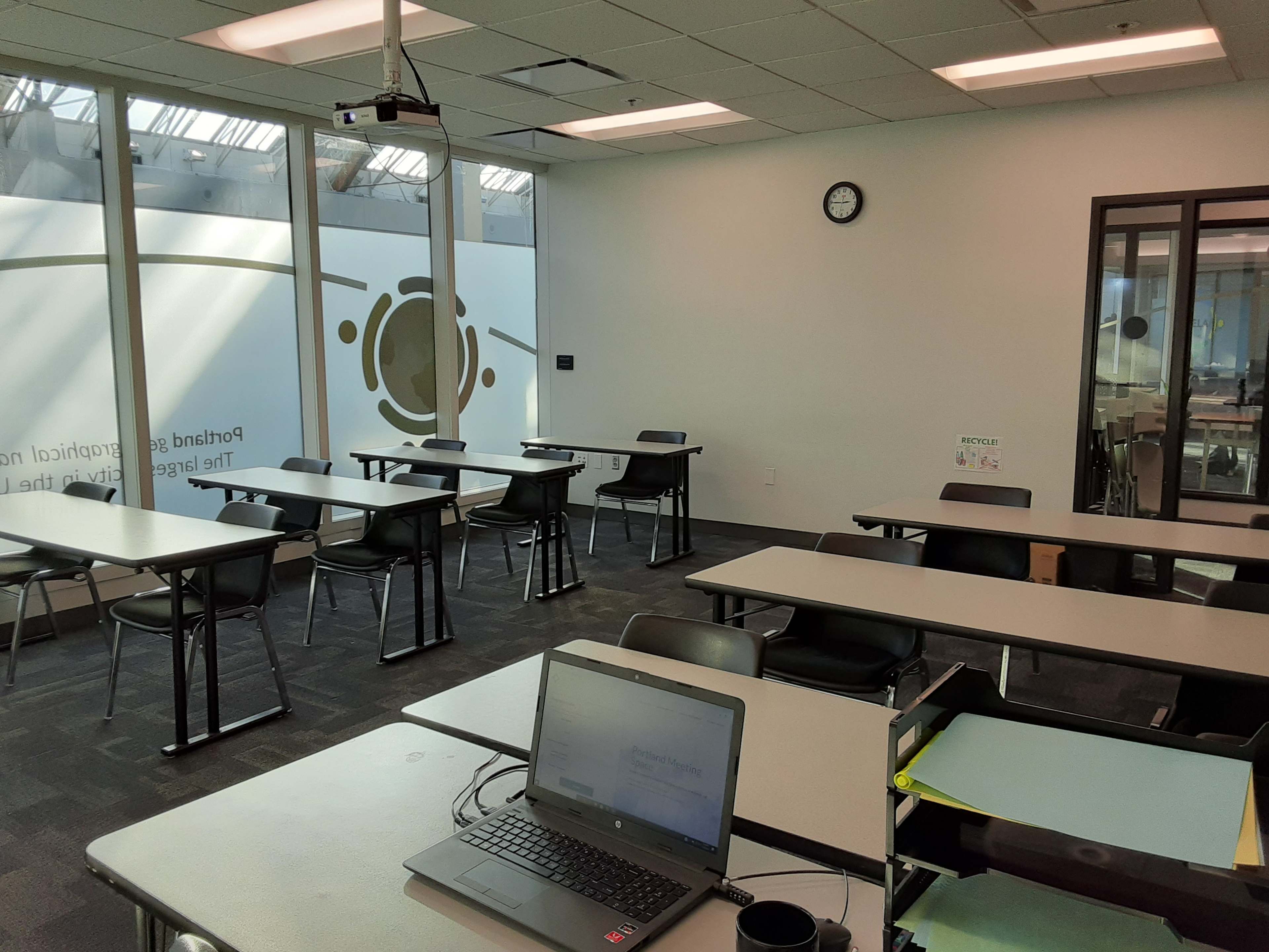 A classroom setting with desks arranged in rows, a laptop on a table, and large windows allowing natural light.