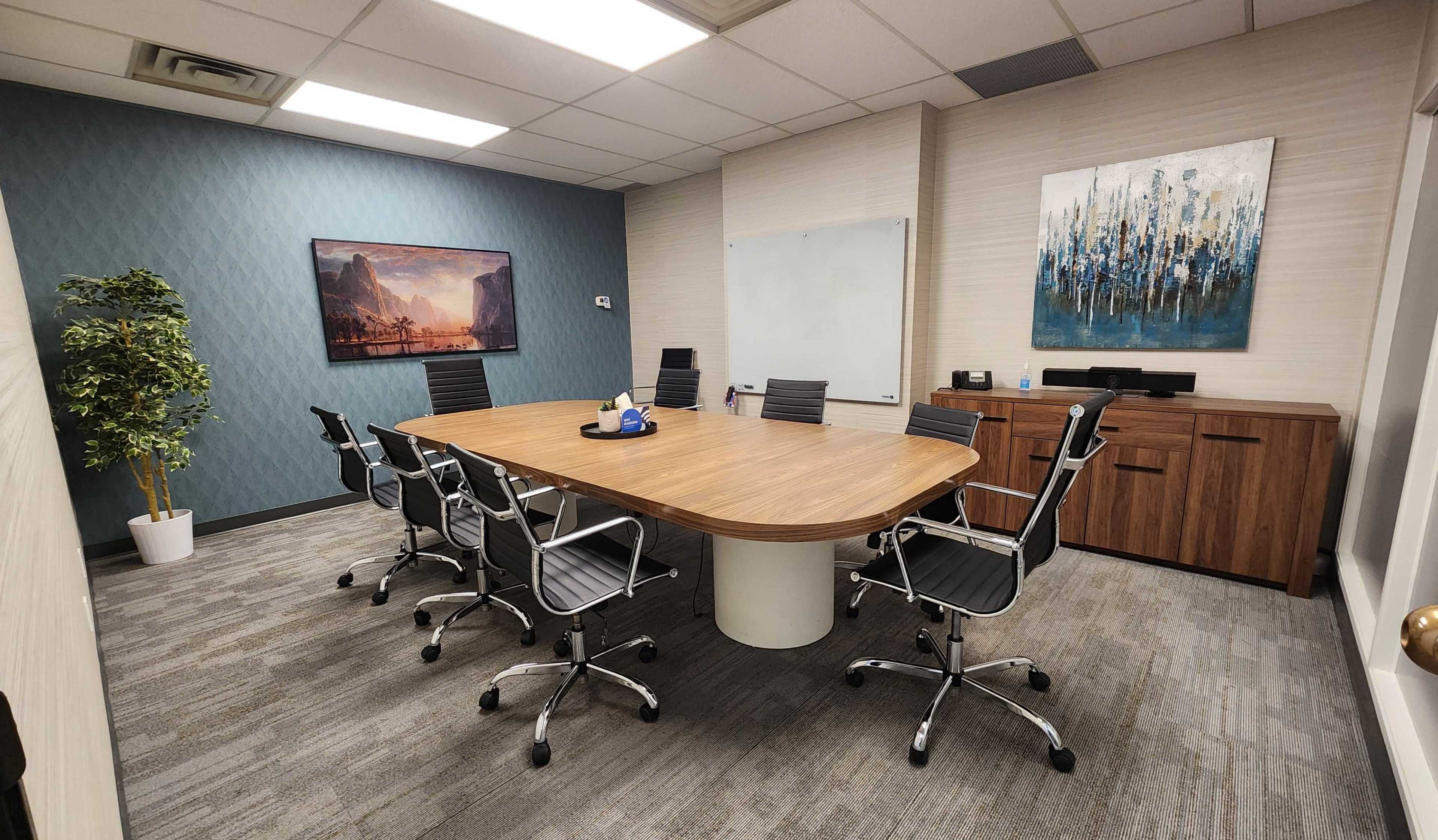 The image shows a modern conference room with an oval wooden table surrounded by six ergonomic chairs, a whiteboard, and wall art.
