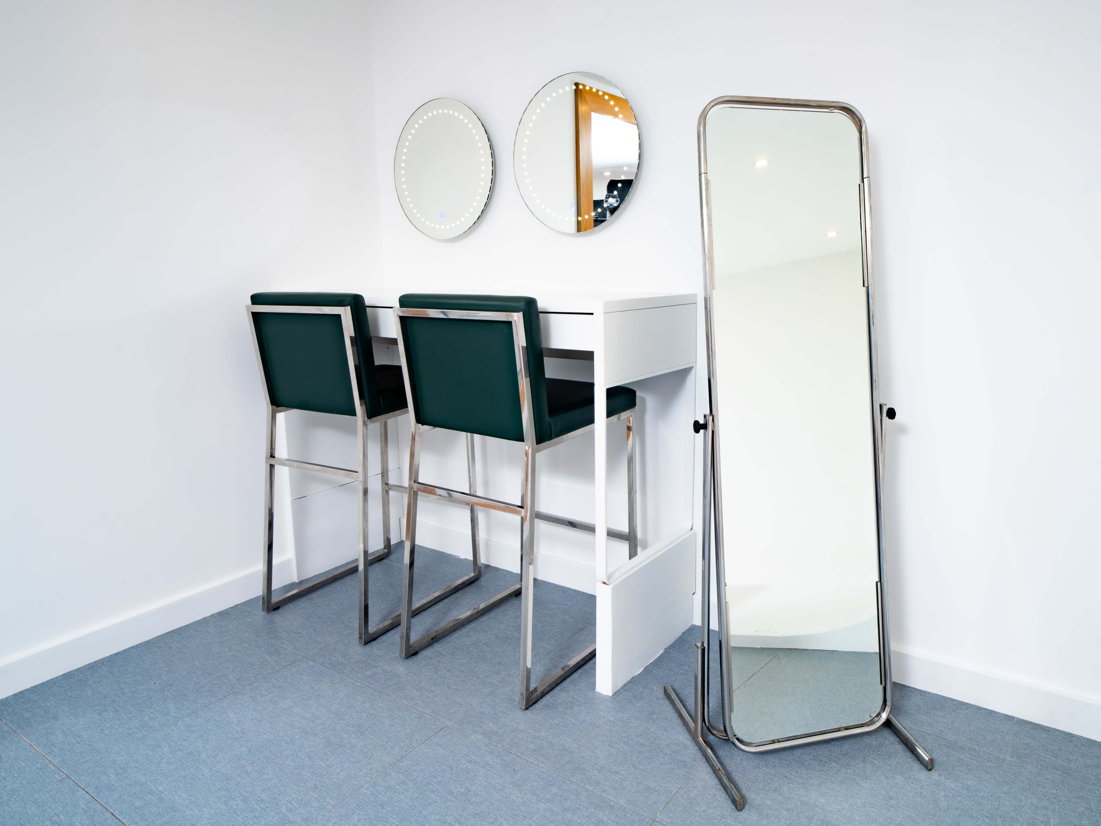 The image shows a minimalist room featuring two high-backed bar stools at a white table, flanked by two circular mirrors and a tall, standing mirror against the wall.
