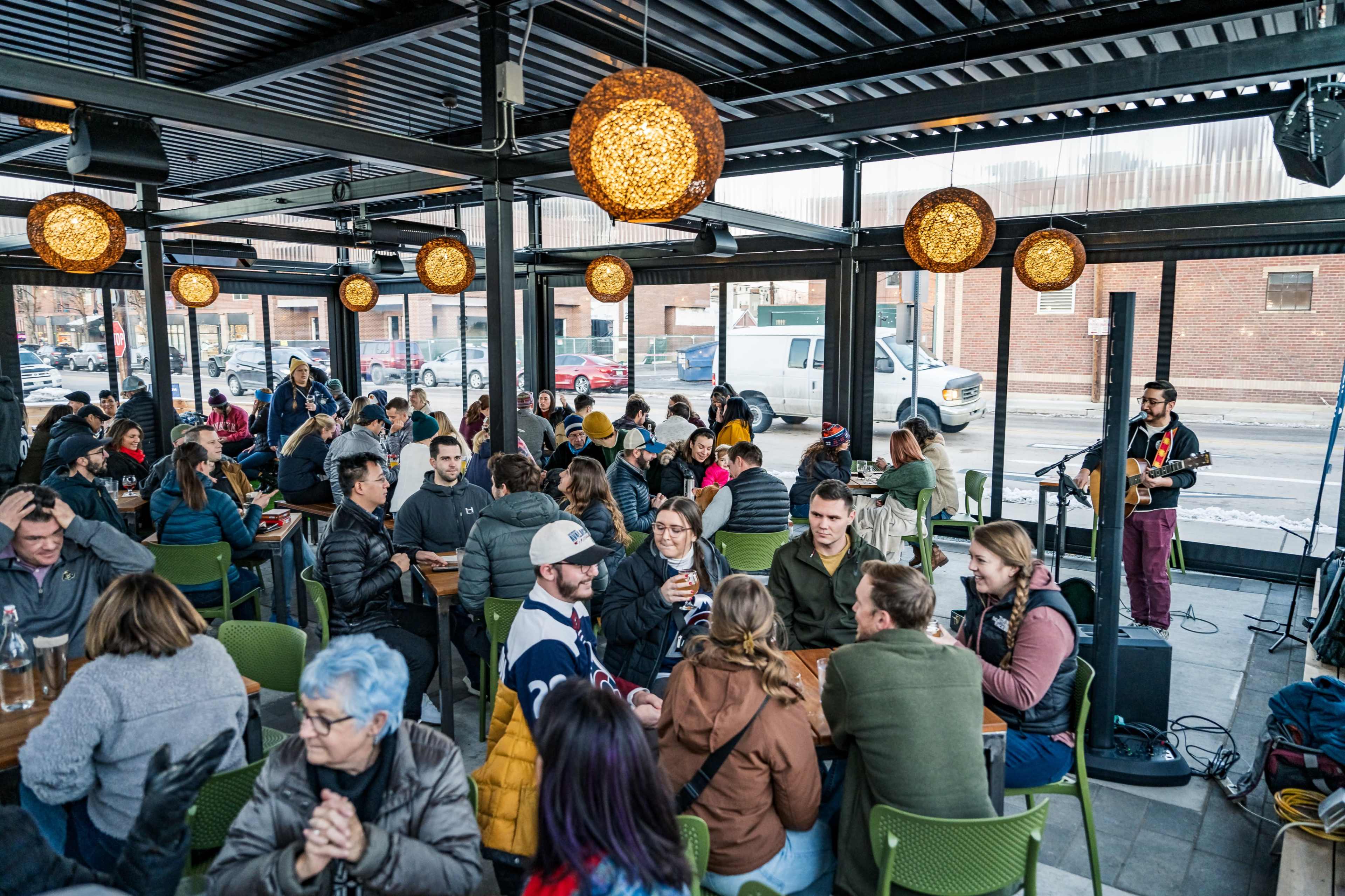 A bustling indoor space with a live musician performing, surrounded by groups of people enjoying drinks and conversation.