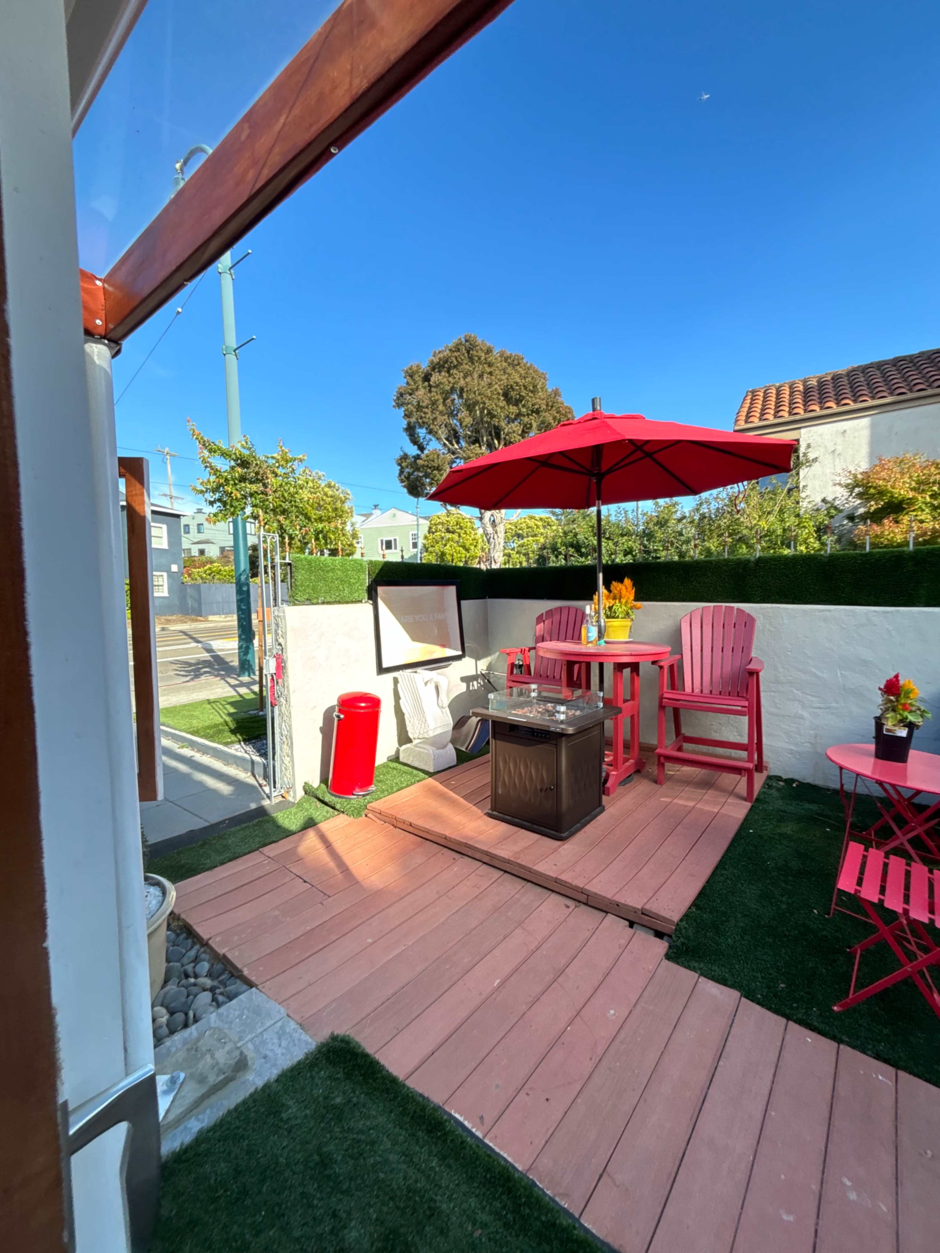 The image shows a small outdoor patio with wooden decking, a red umbrella, pink chairs, a table with a floral arrangement, and a small fire pit surrounded by green grass and hedges.