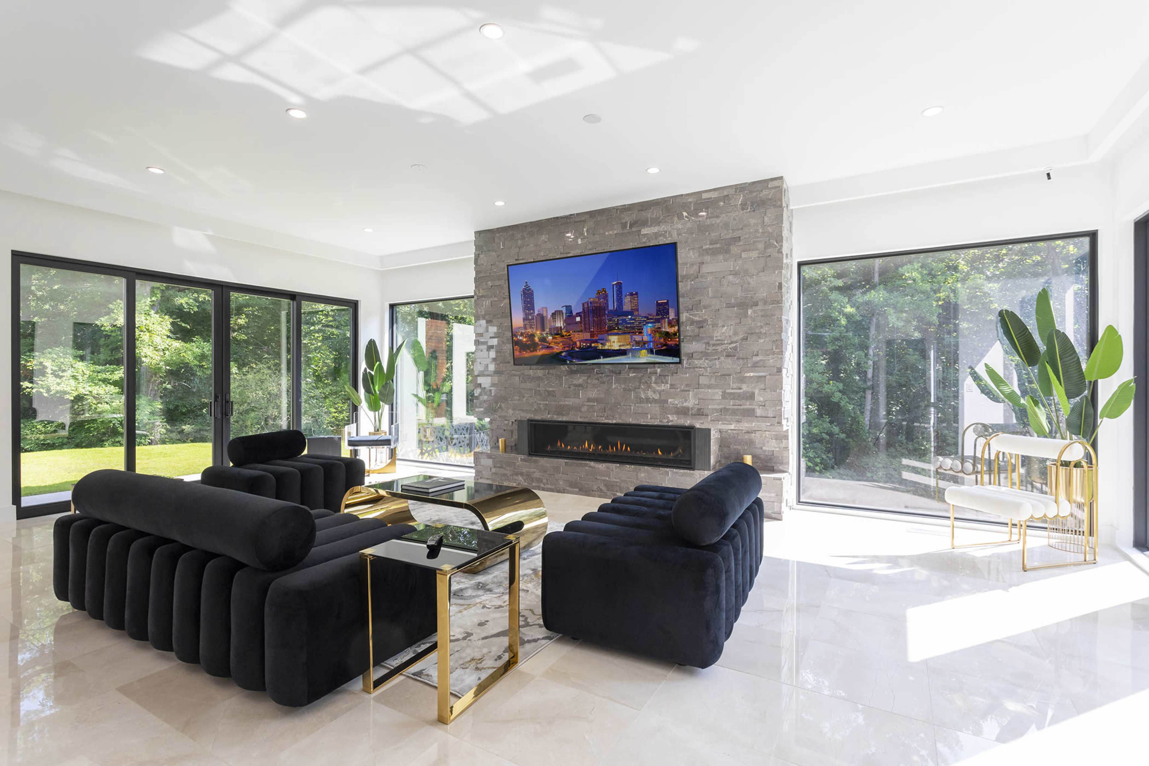 The spacious living room features black sofas, a stone fireplace, a large television, and windows overlooking a green outdoor area.