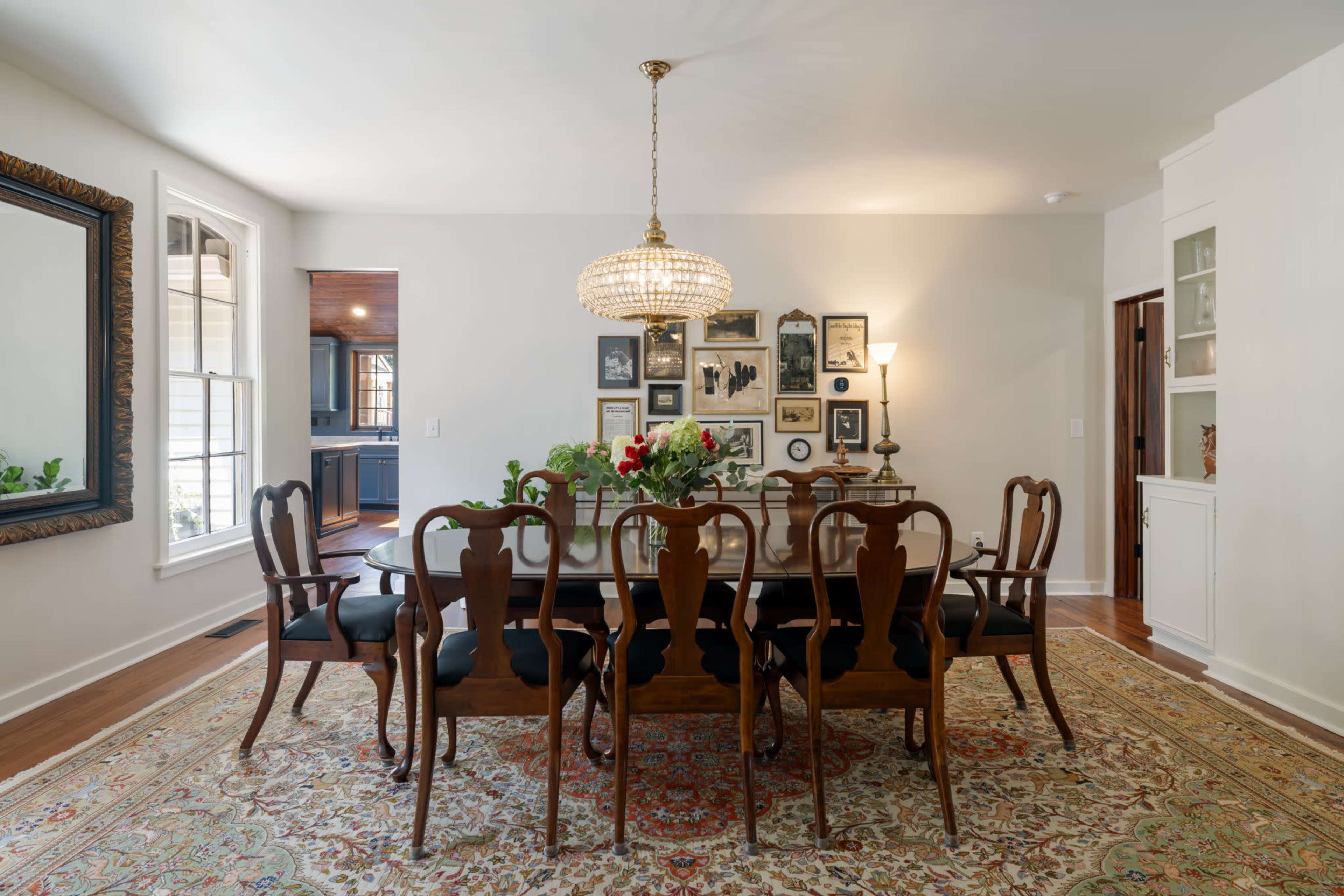 A dining room features a long wooden table surrounded by elegant chairs, a chandelier overhead, and a gallery wall adorned with framed pictures.
