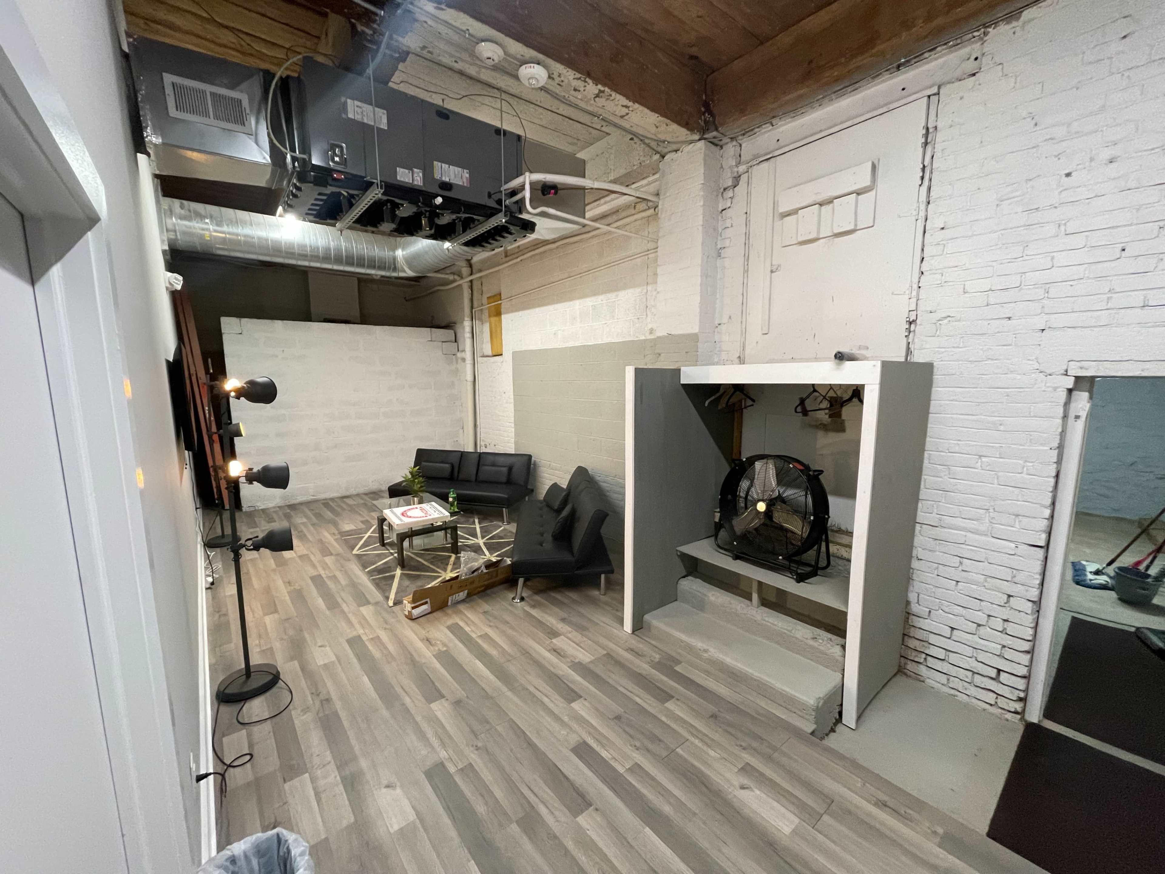 The image shows a modern, minimalist interior space featuring a black sofa, a coffee table, and a clothing rack near a wall, with exposed ductwork and wooden beams overhead.