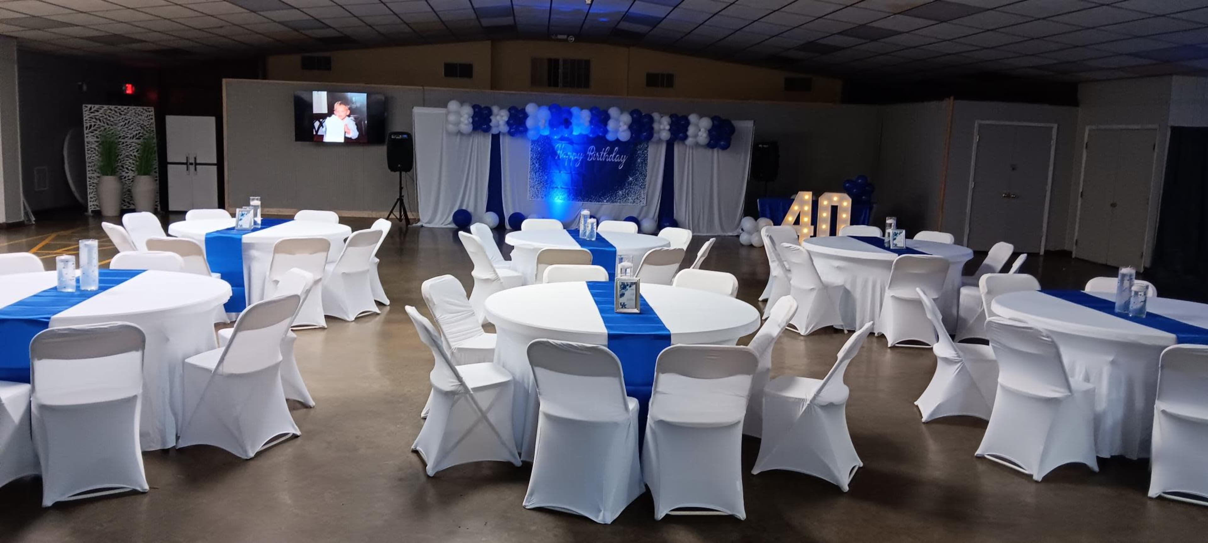 The image shows a decorated event space with round tables covered in white tablecloths and blue accents, arranged for a gathering, and featuring a backdrop with balloons and a screen displaying a video.