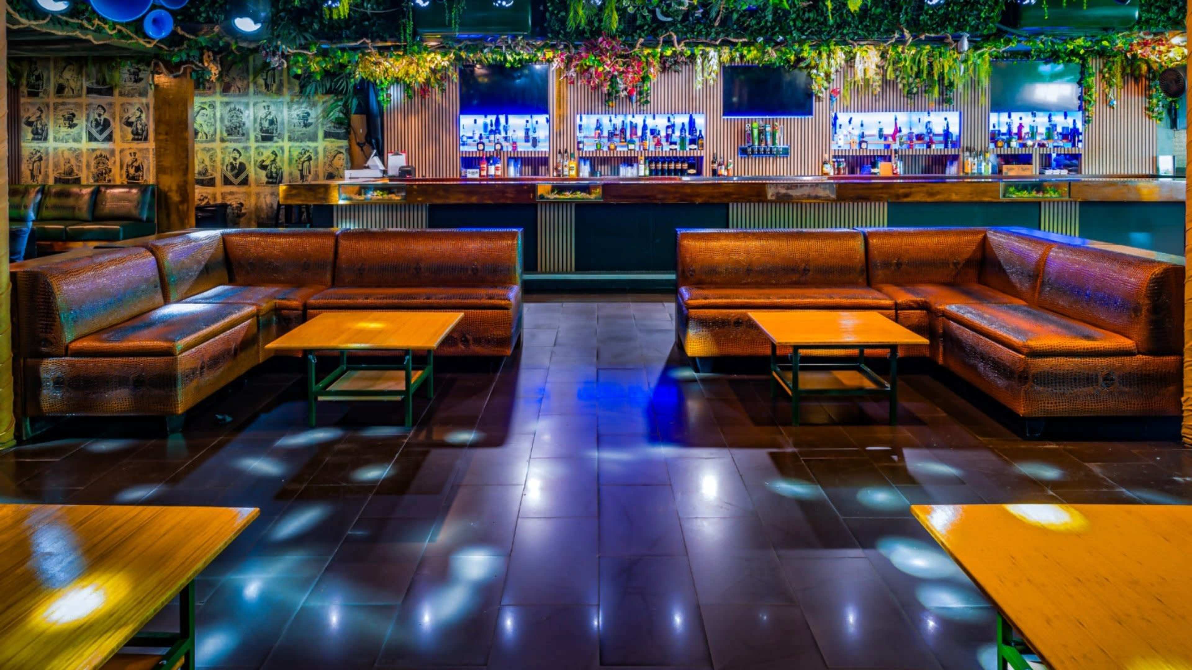 A modern lounge area with brown leather sofas and wooden tables, illuminated by colorful lights and surrounded by greenery, with a bar area in the background.