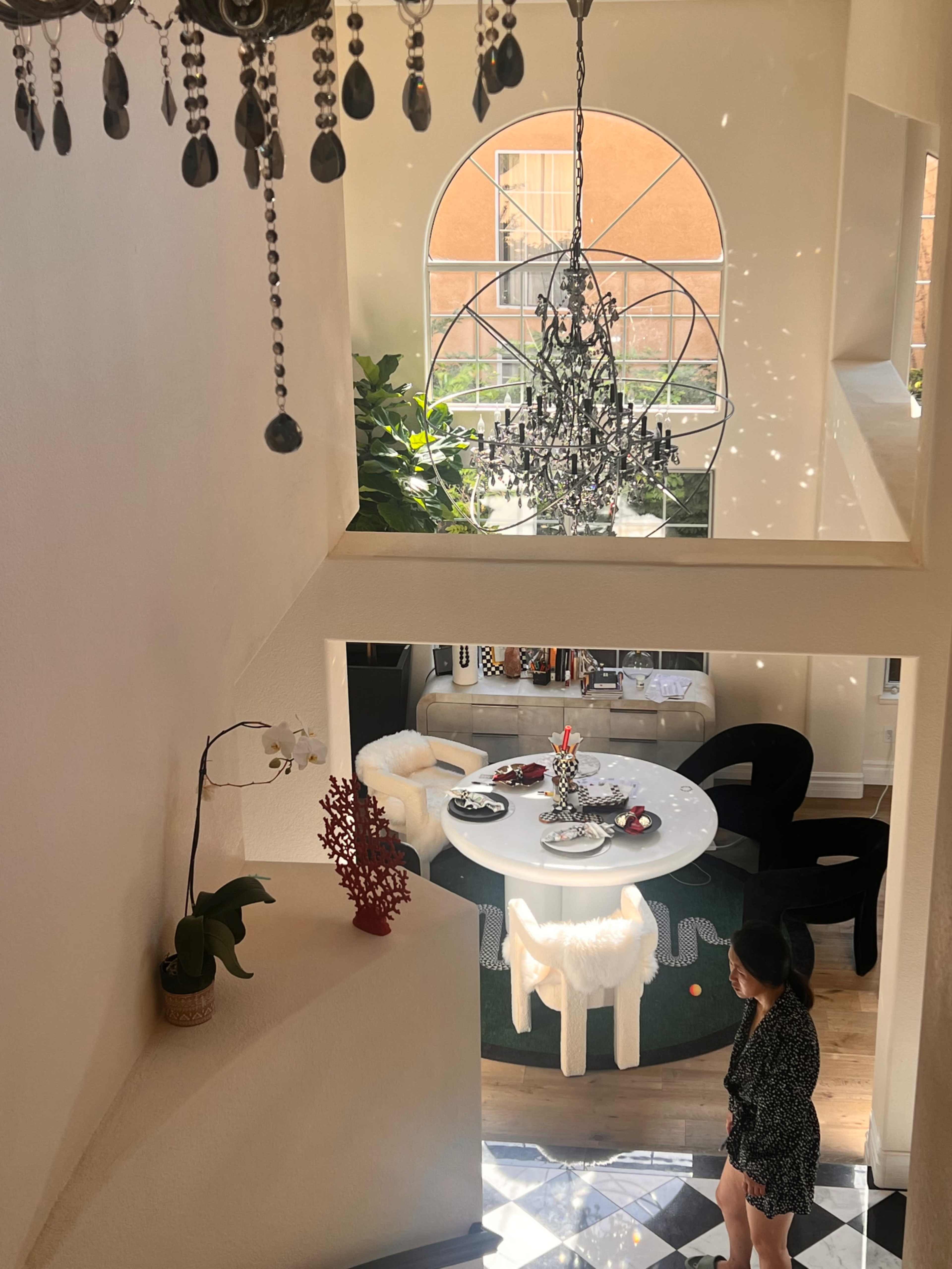 A woman stands at the top of a stairway overlooking a dining area set with a table decorated for a meal, featuring a chandelier above and large windows allowing natural light.