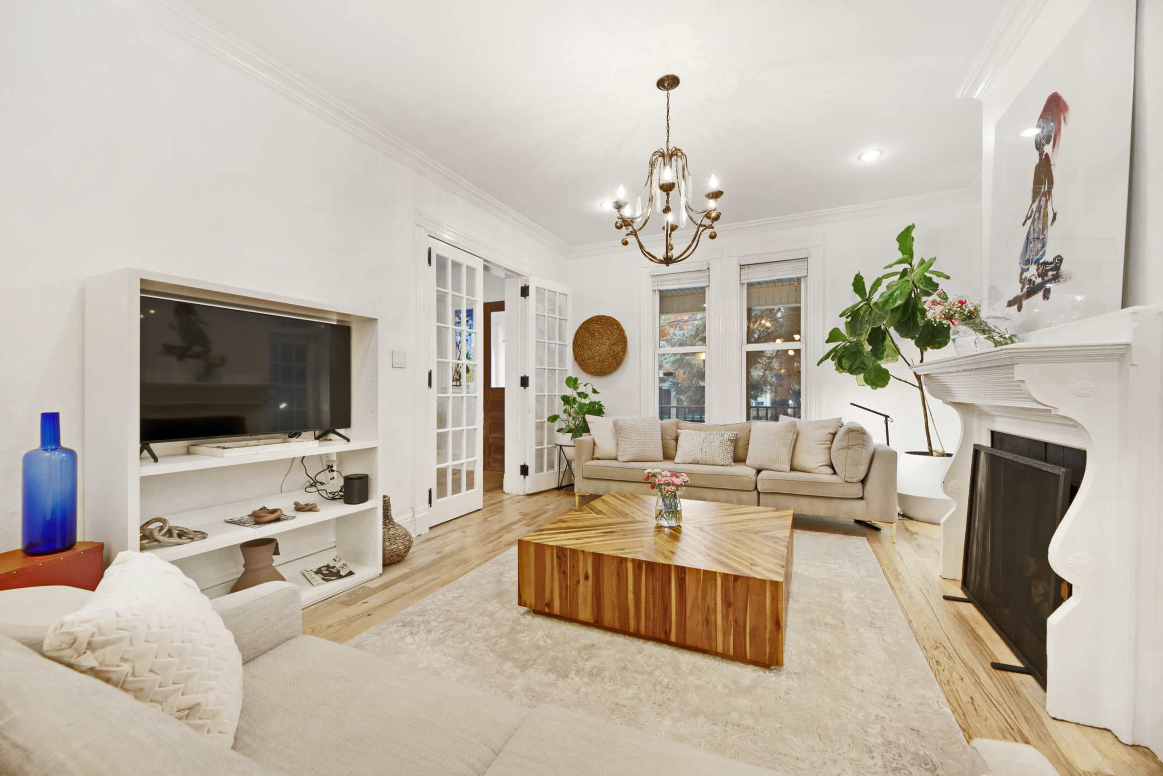 A modern living room features a beige sectional sofa, a wooden coffee table, a television mounted on a white shelf, and a fireplace surrounded by large windows and indoor plants.