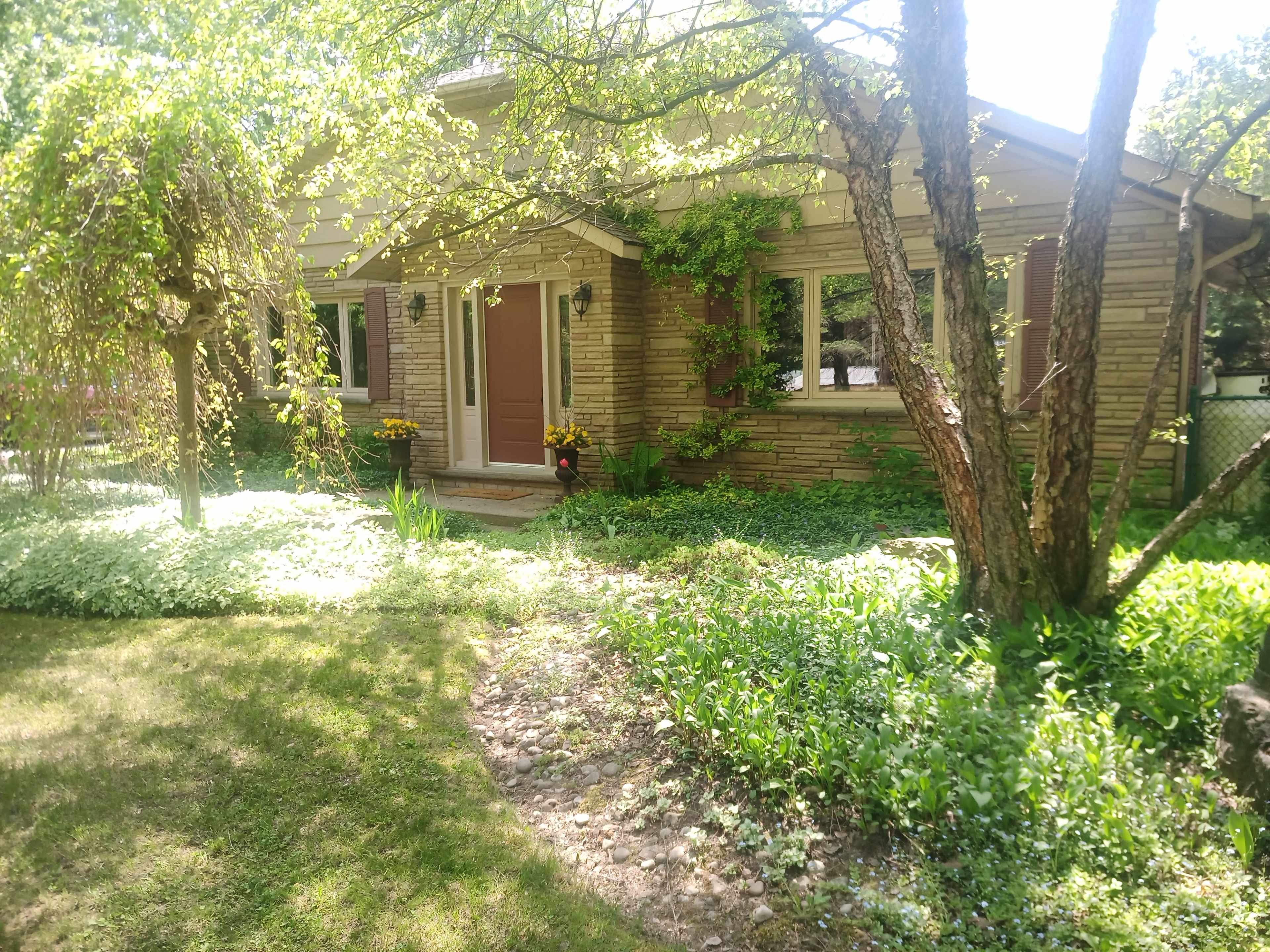 A single-story house with brick siding, a front door flanked by two windows, surrounded by a landscaped yard featuring green grass and flower beds.