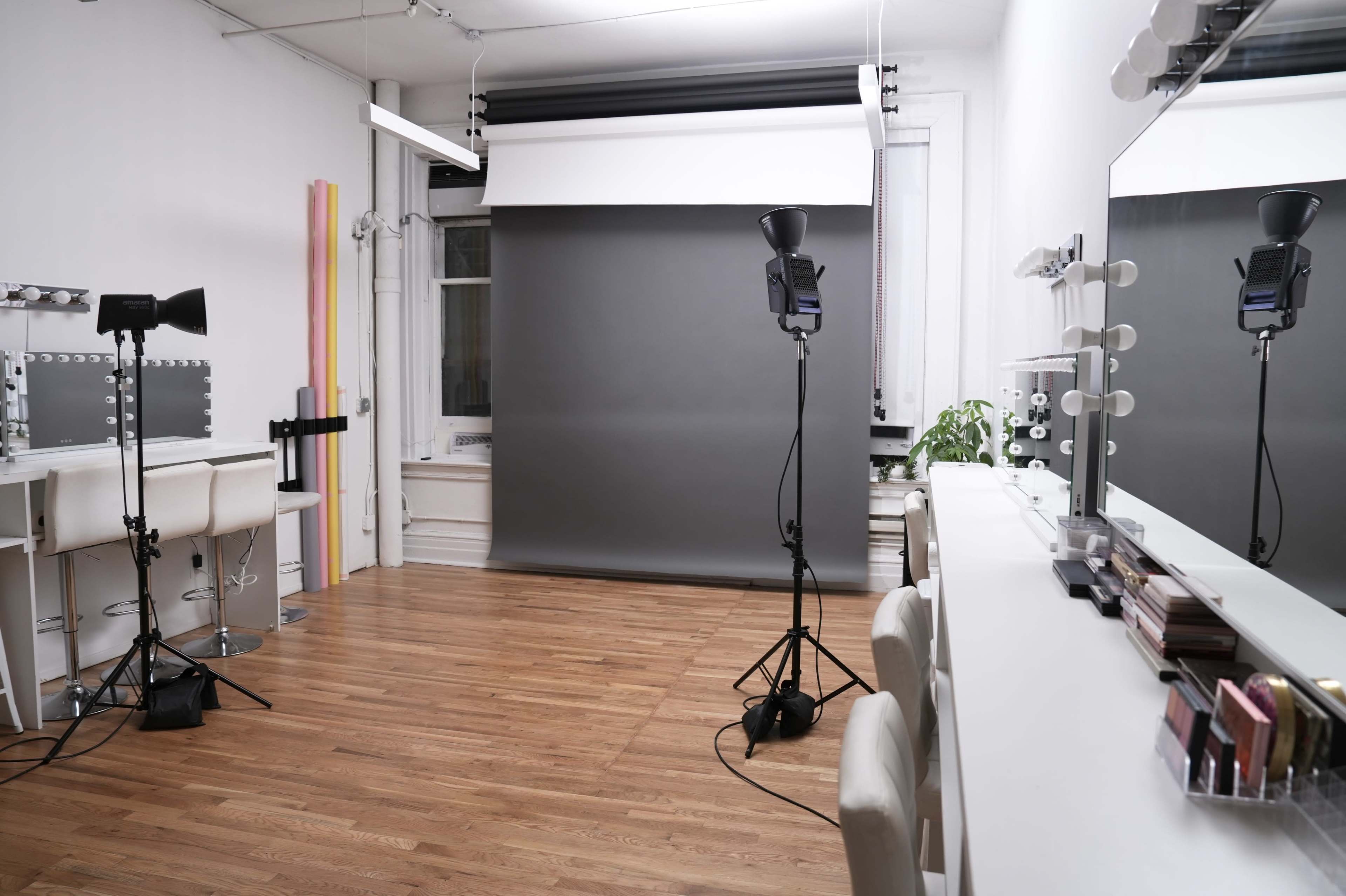The image shows a well-lit photography studio with a gray backdrop, two light stands, and a vanity area featuring mirrors and makeup products.