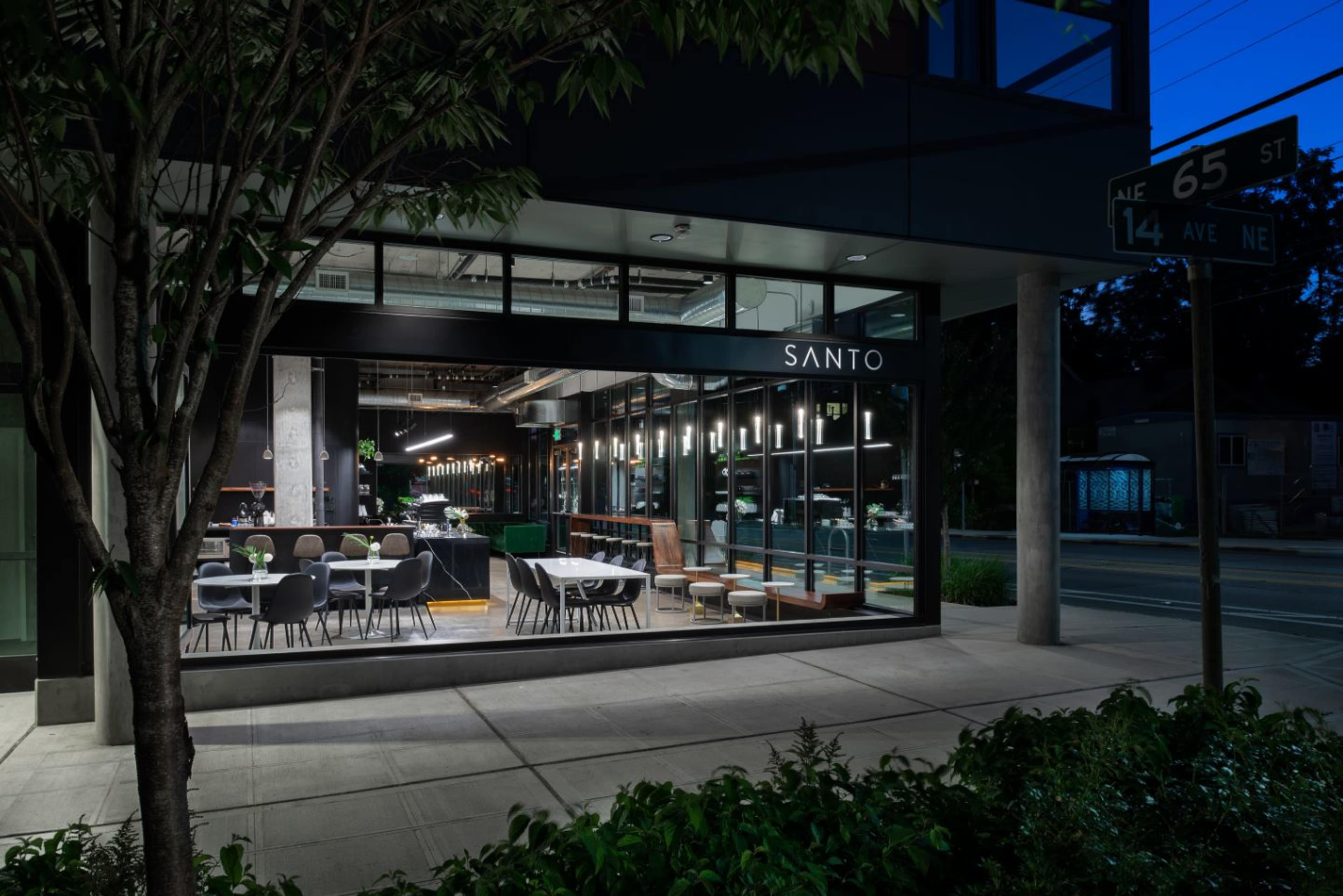 The image shows a restaurant named "Santo" with large glass windows, featuring a dining area illuminated at night, located on a street corner.