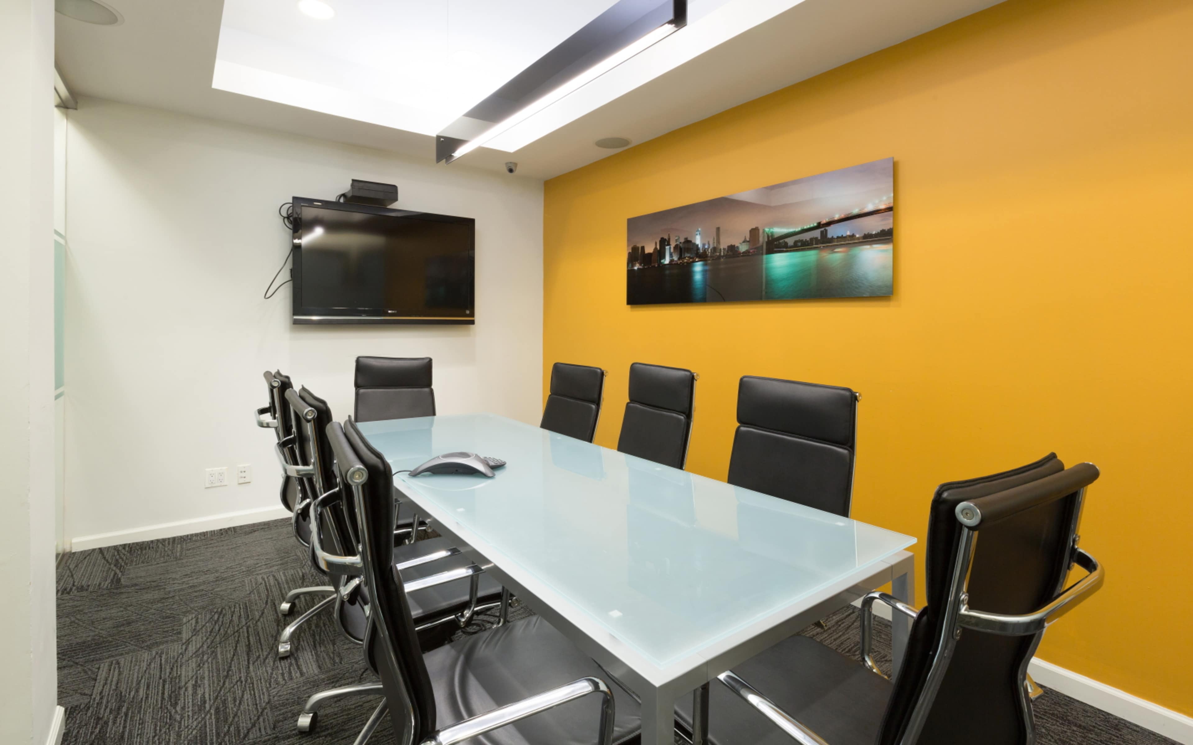 A conference room features a glass table surrounded by black chairs, with a wall-mounted TV and a vibrant orange accent wall.