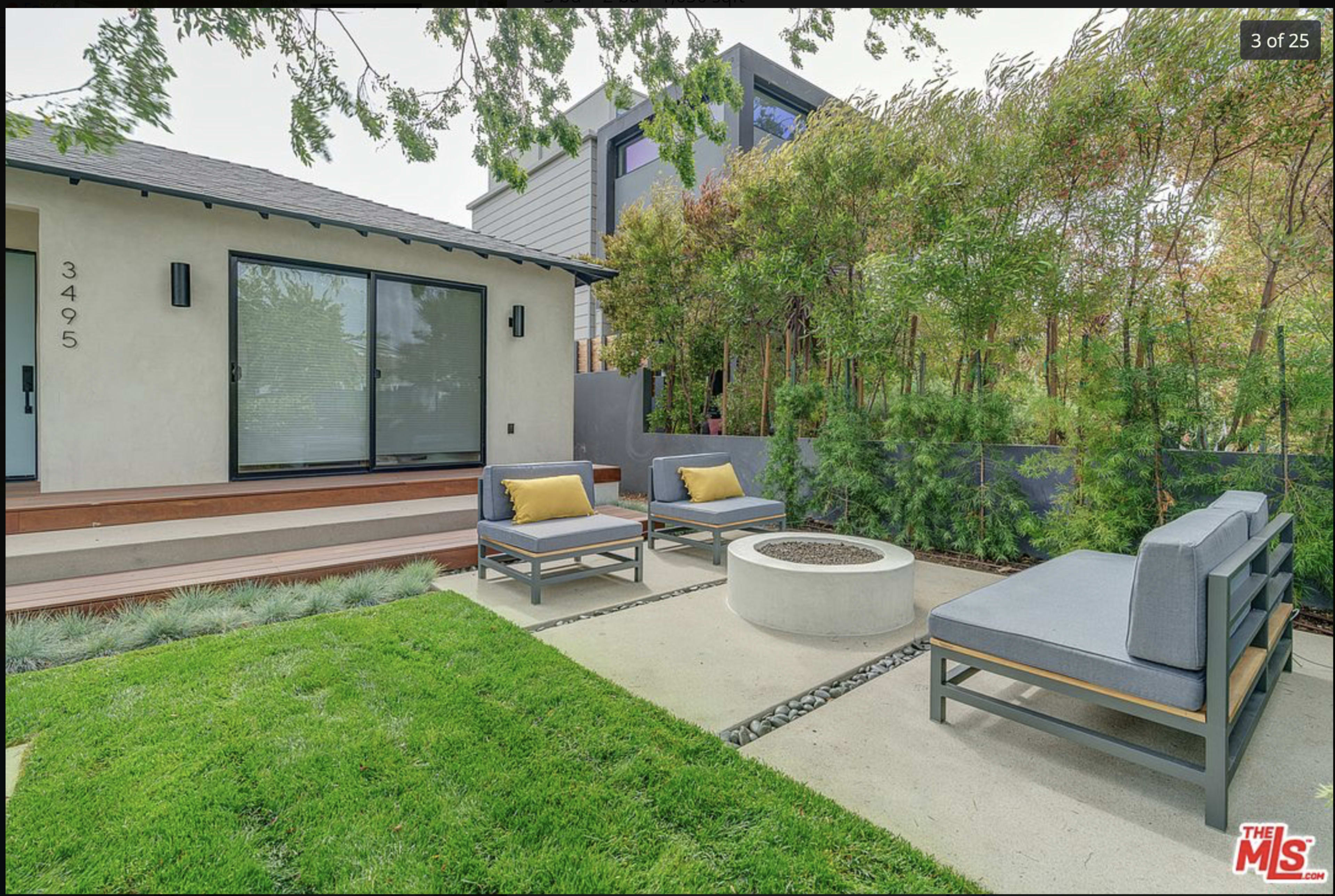 A modern outdoor space features two gray seating areas with yellow cushions arranged around a circular fire pit, alongside lush green grass and bamboo landscaping.