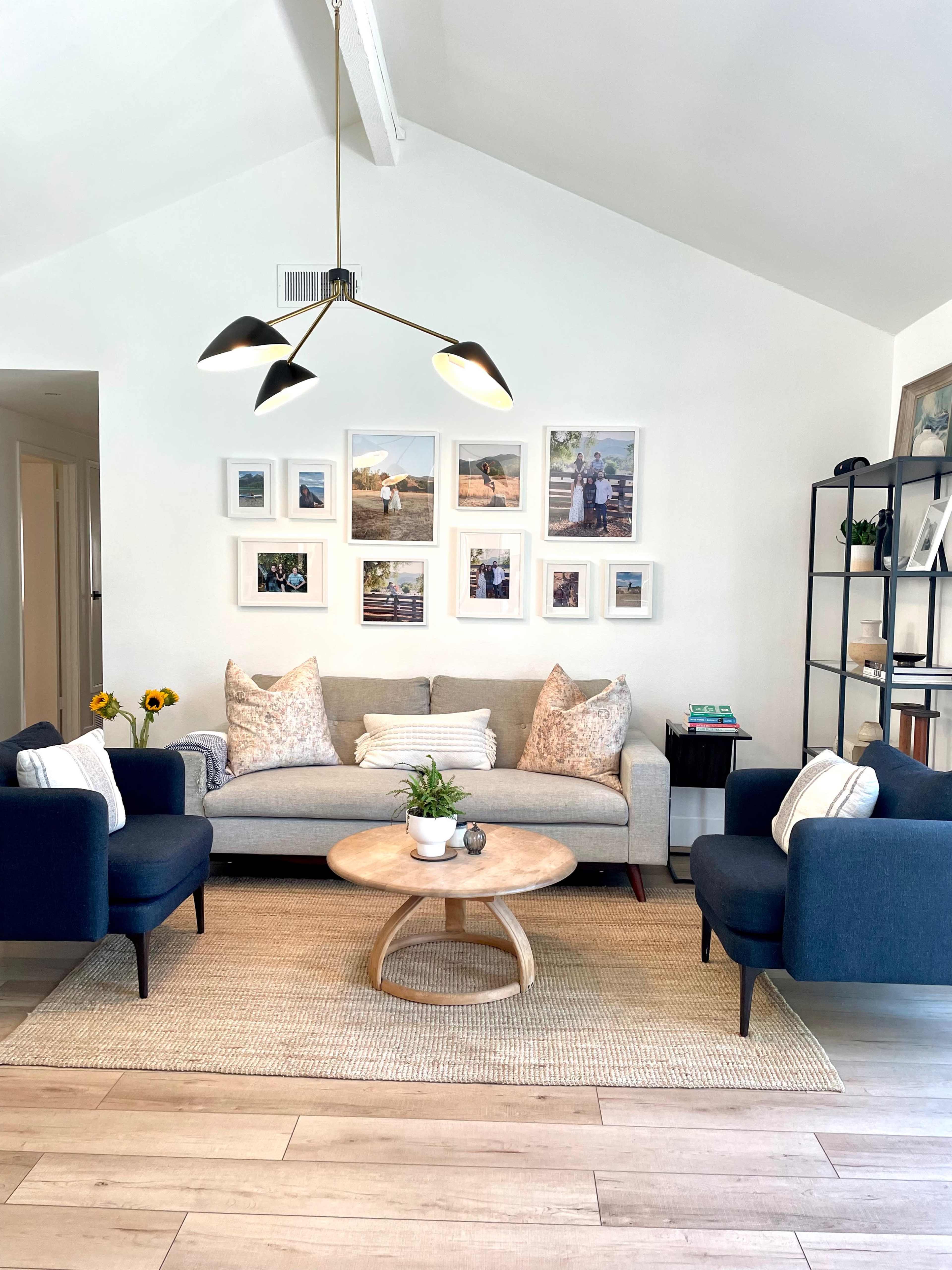 A bright living room features a gray sofa with decorative pillows, surrounded by two blue chairs and a circular coffee table, with framed photos hanging on the white wall.