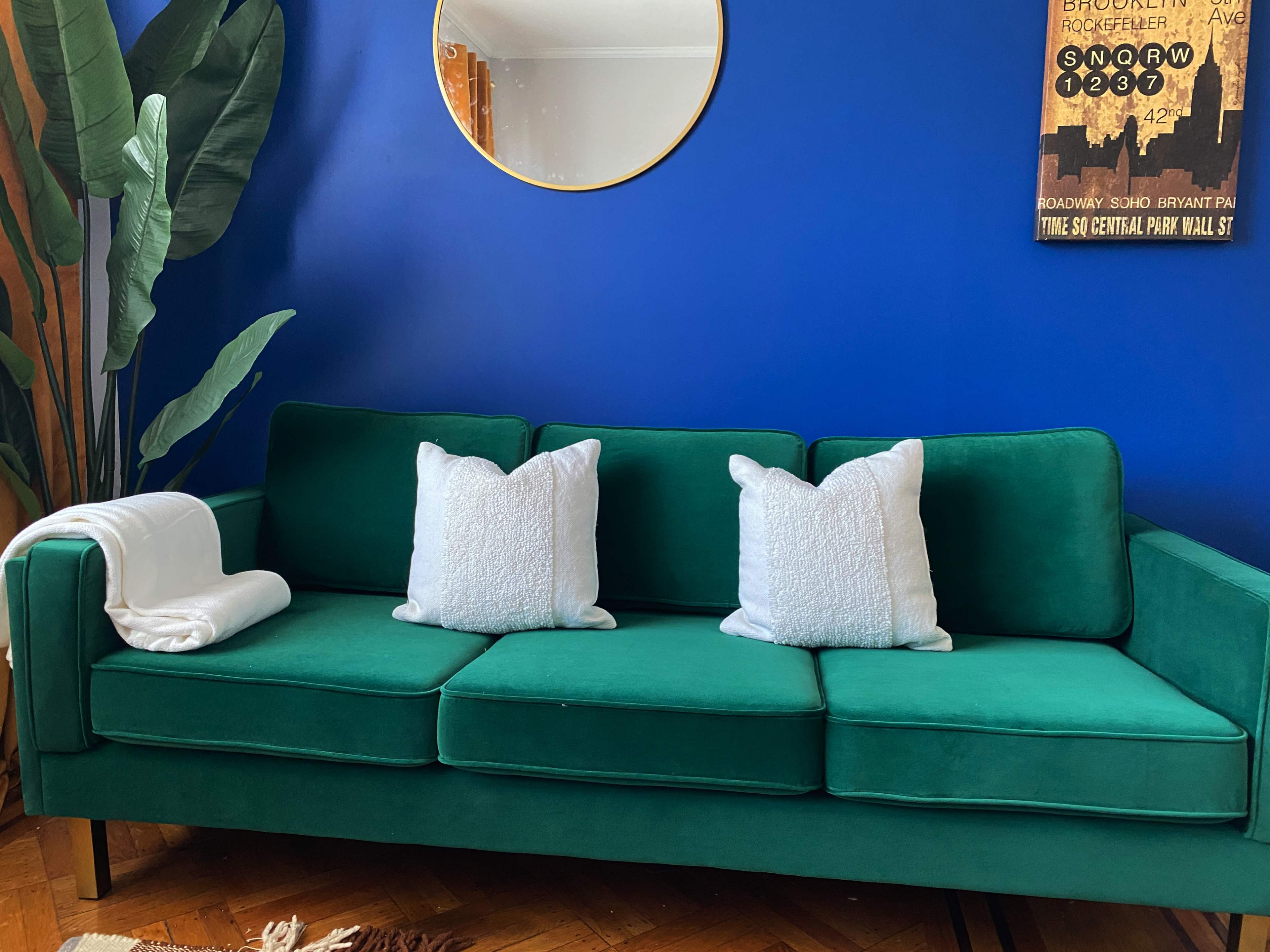 A green sofa with two white pillows, placed against a blue wall with a round mirror and a framed poster.