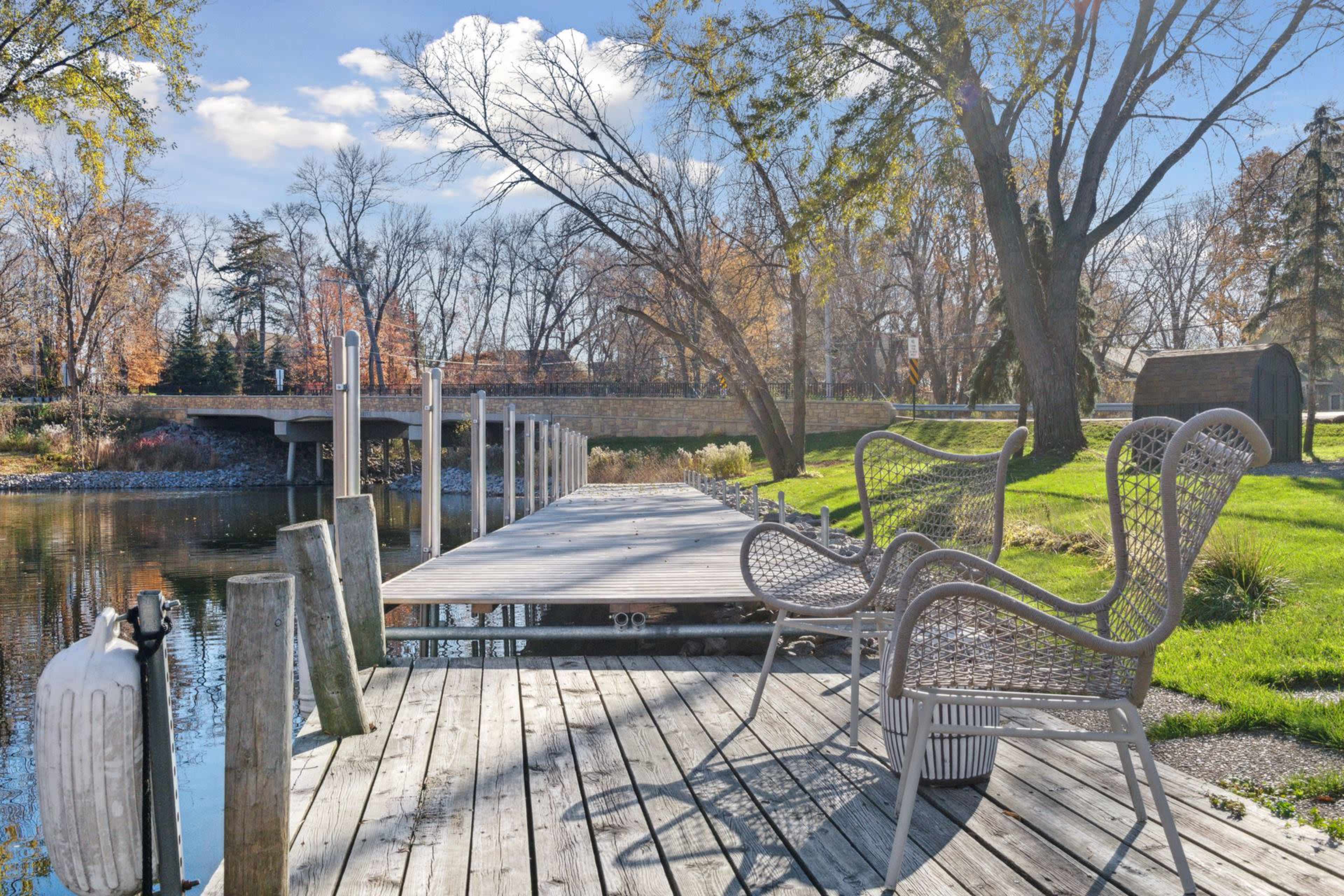 A wooden dock extends over a calm waterway, lined with chairs and surrounded by trees and a bridge in the background.