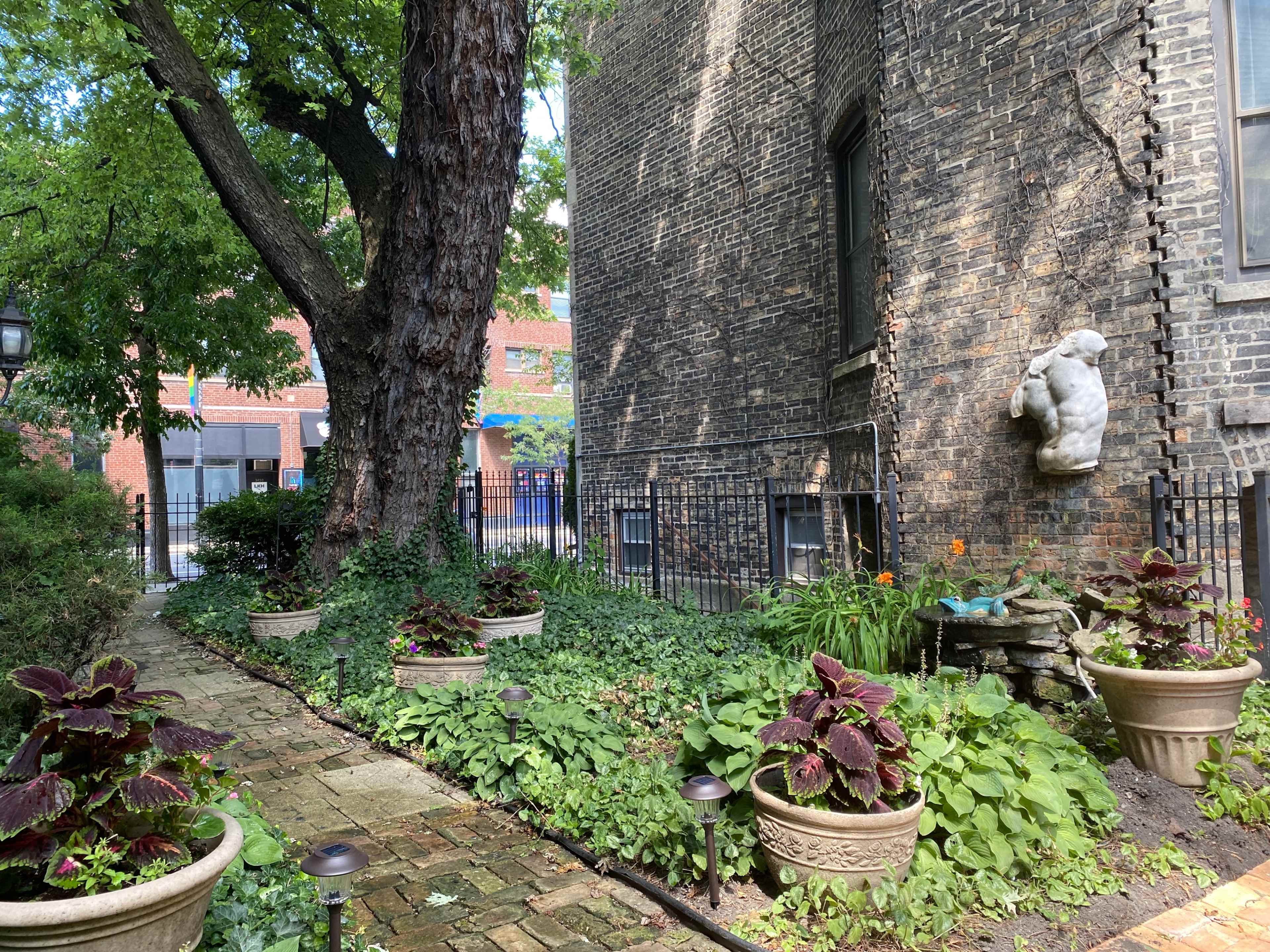 A stone sculpture of a bear is mounted on a brick wall adjacent to a lush, landscaped garden filled with potted plants and a winding stone path.