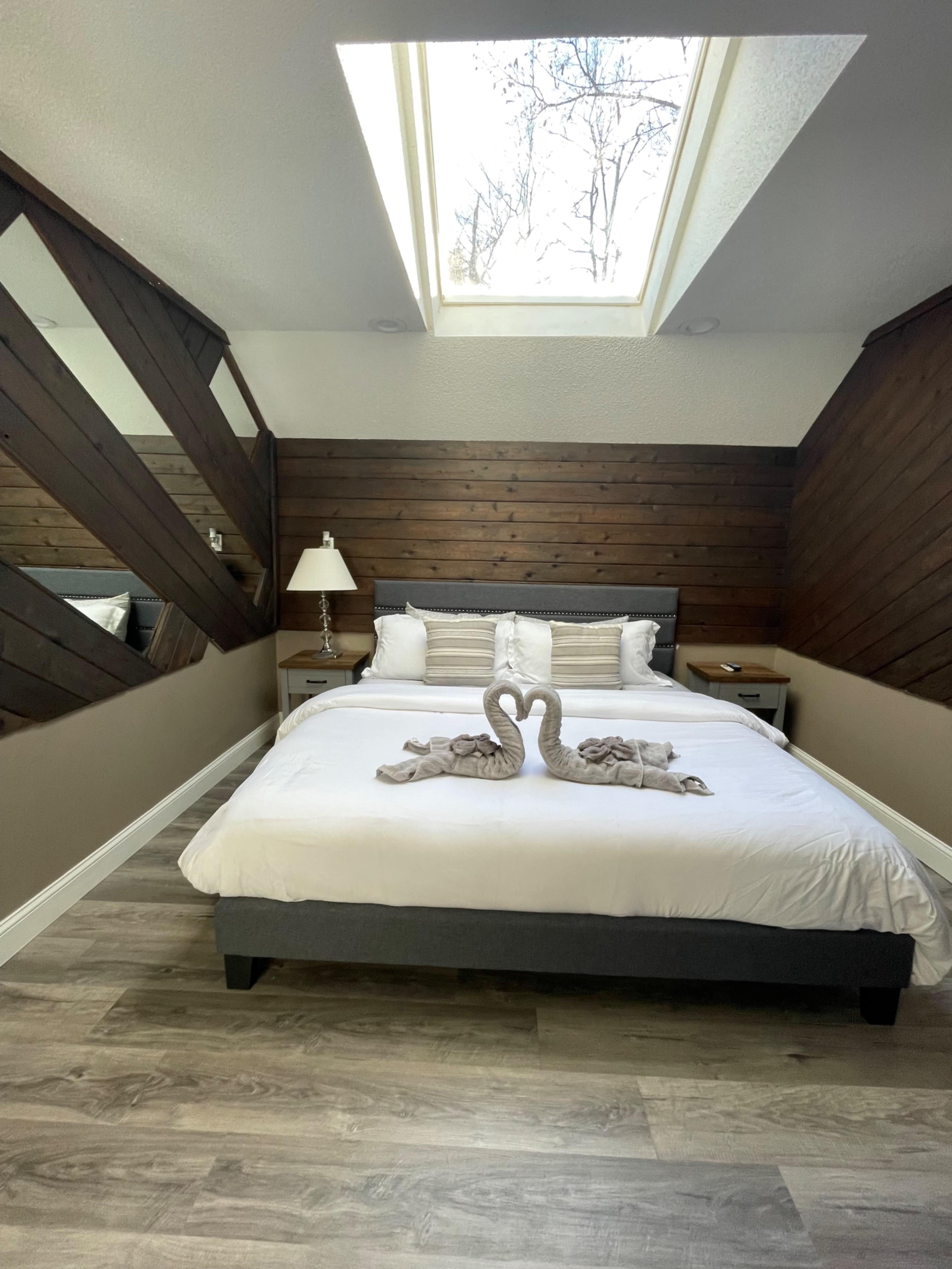 A bedroom features a skylight above a neatly made bed with a decorative towel swan at its foot and wooden walls.