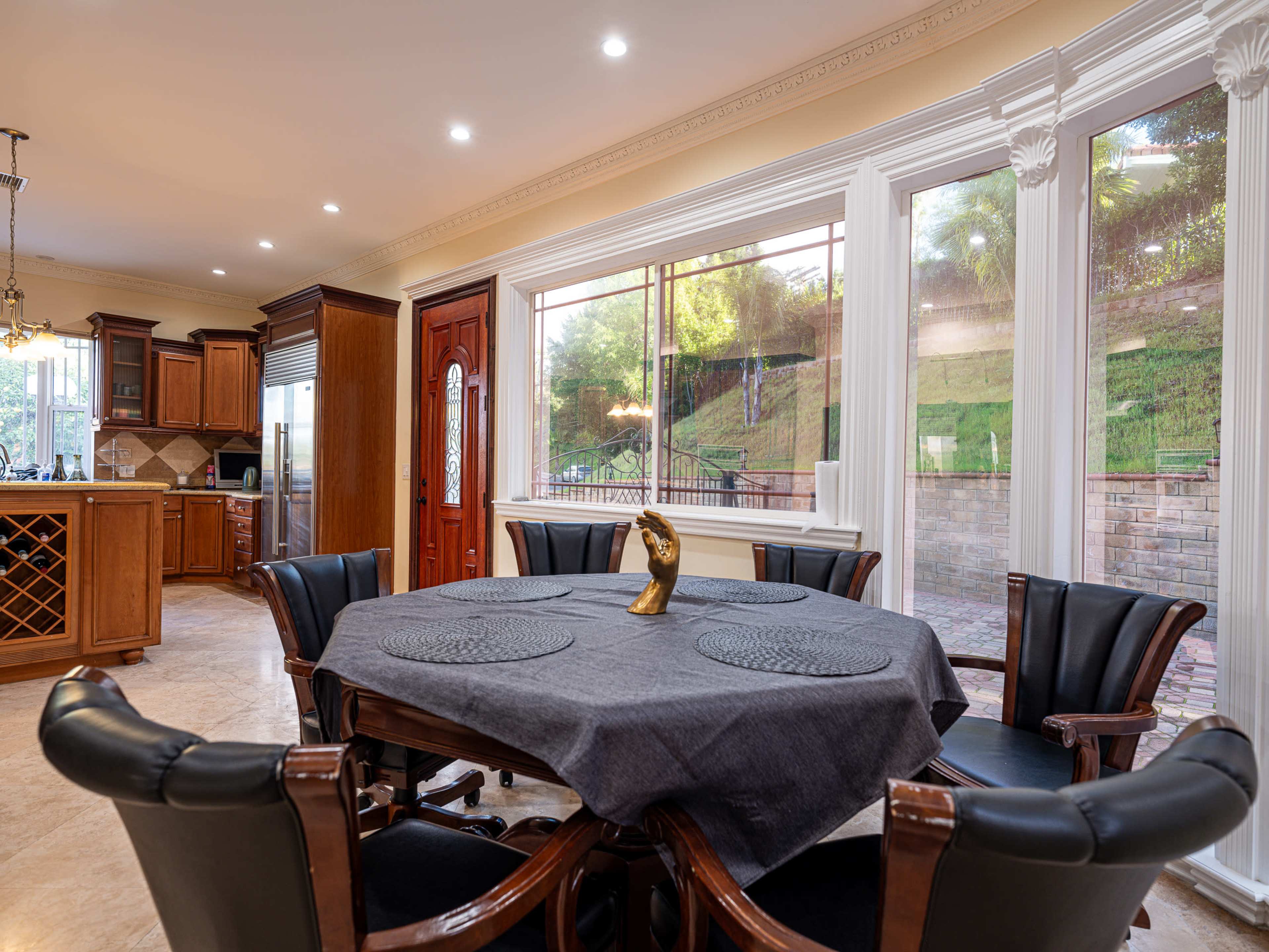 A dining area features a round table with a gray table cover surrounded by black chairs, with large windows offering a view of a landscaped backyard.
