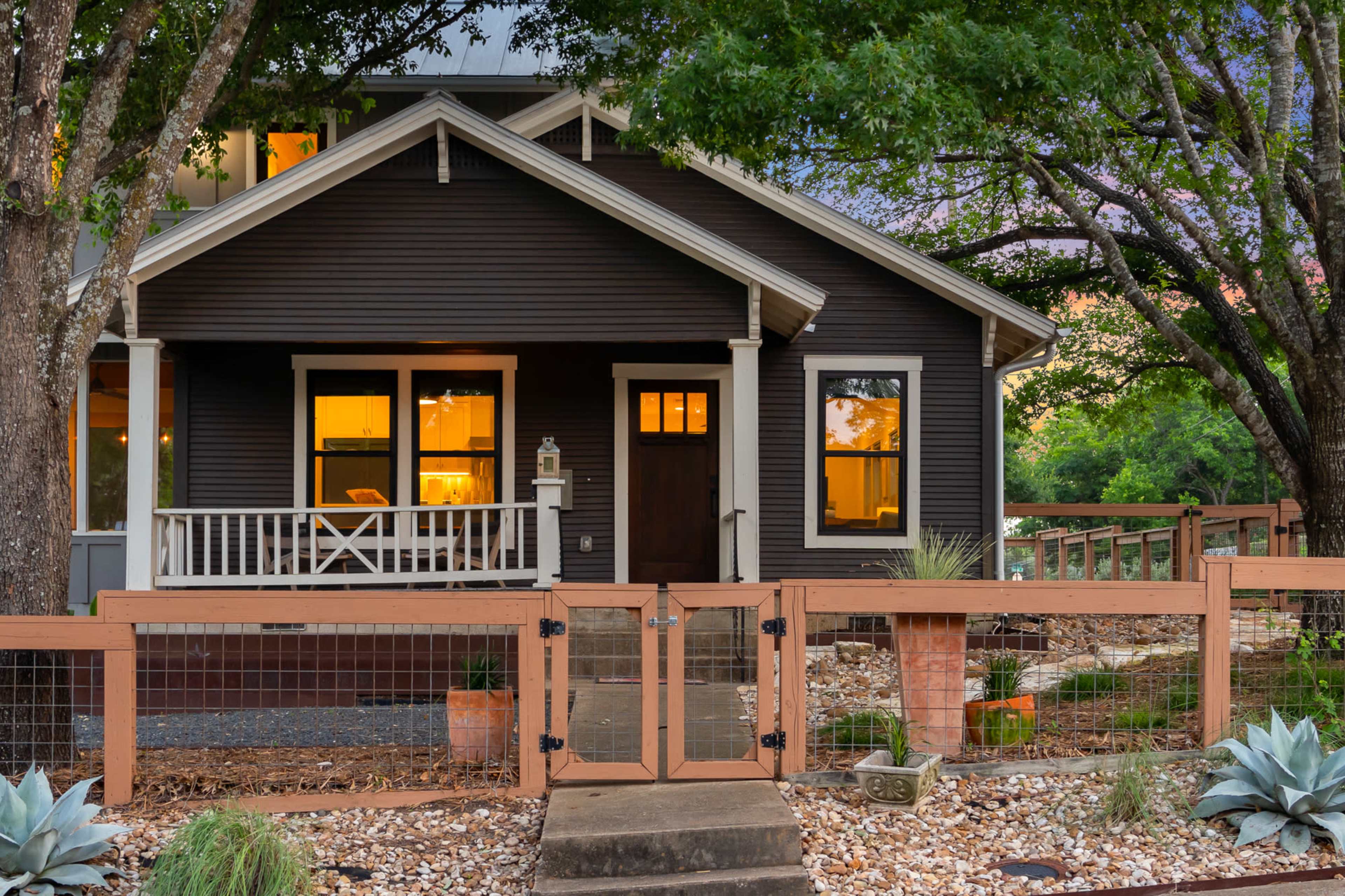 A dark-colored house with a front porch is surrounded by trees and features a fenced yard with garden elements.