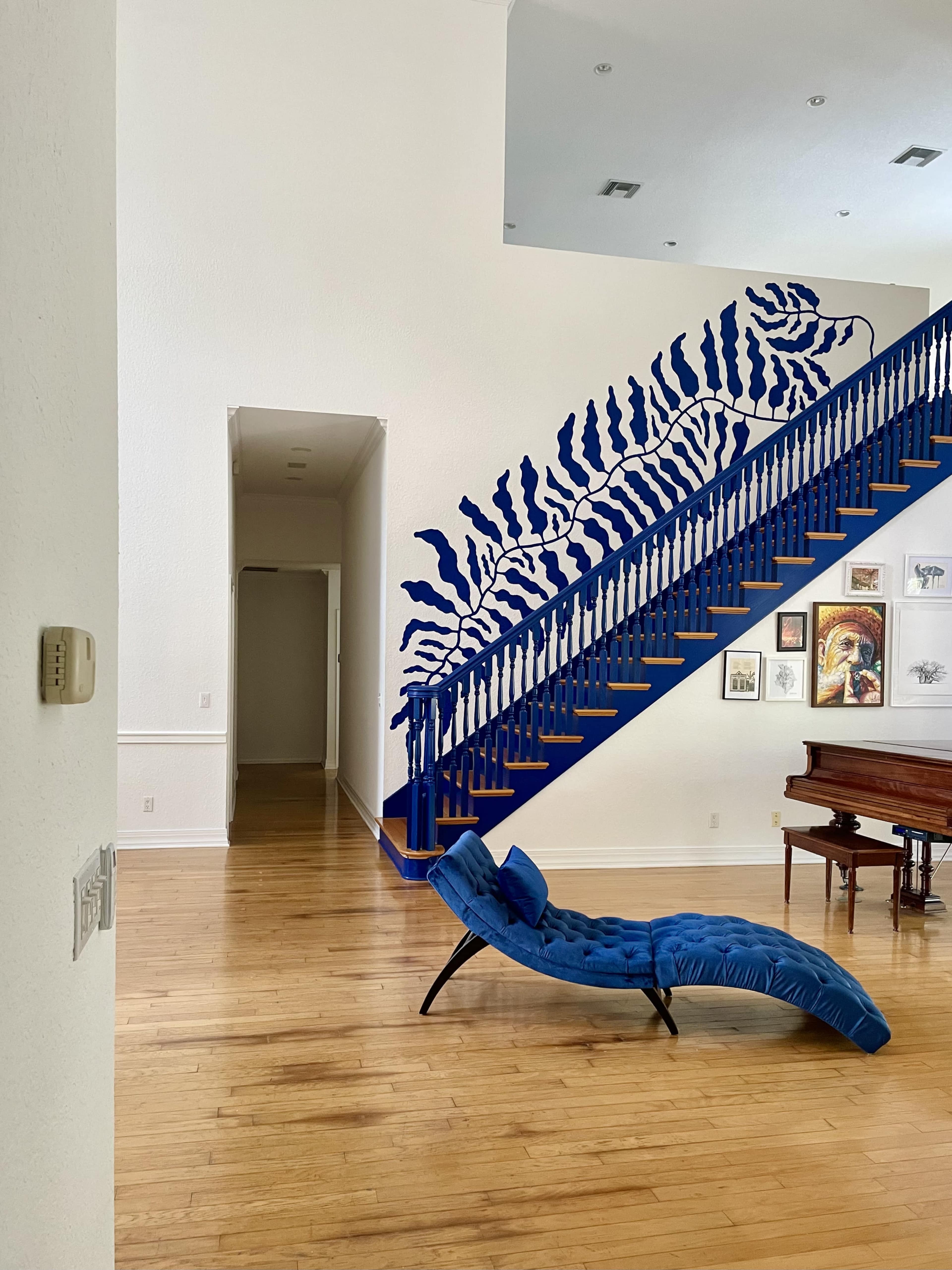The image shows a spacious room with wooden floors featuring a blue staircase adorned with a mural, a blue chaise lounge, and a piano against the wall.