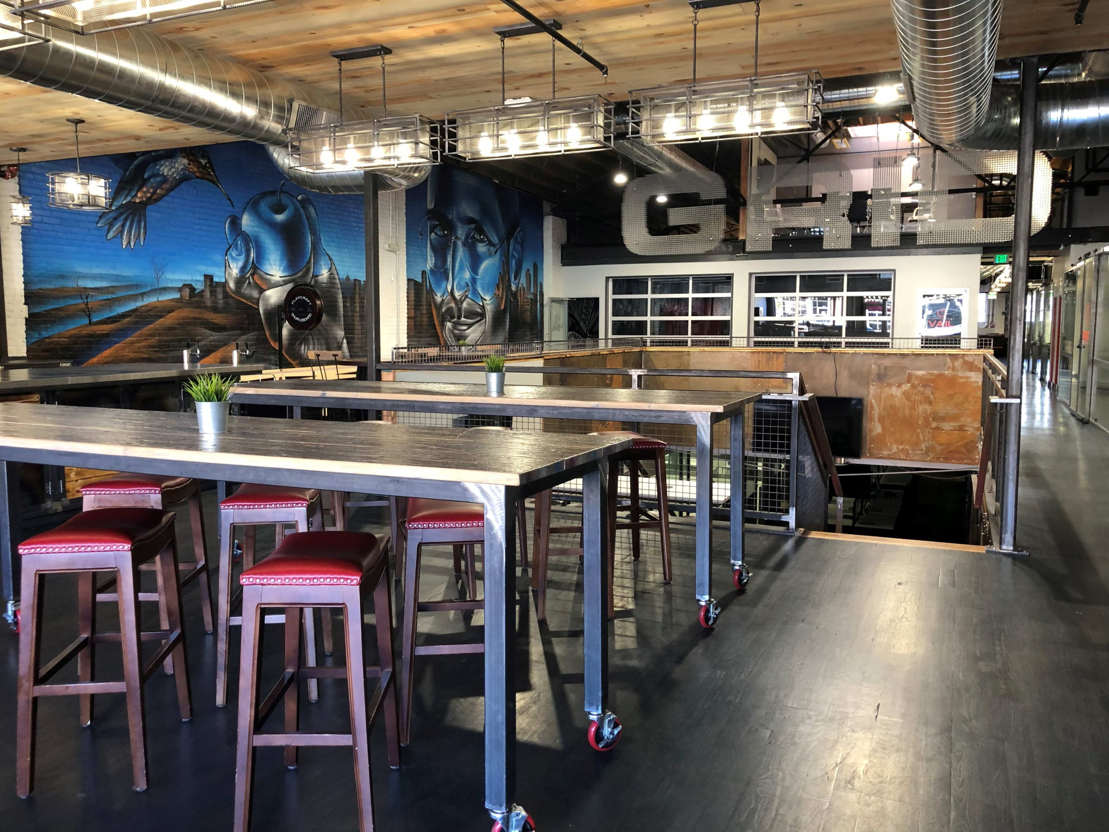 The image shows a modern dining area with high-top tables and bar stools, featuring industrial decor and a large mural on the wall.