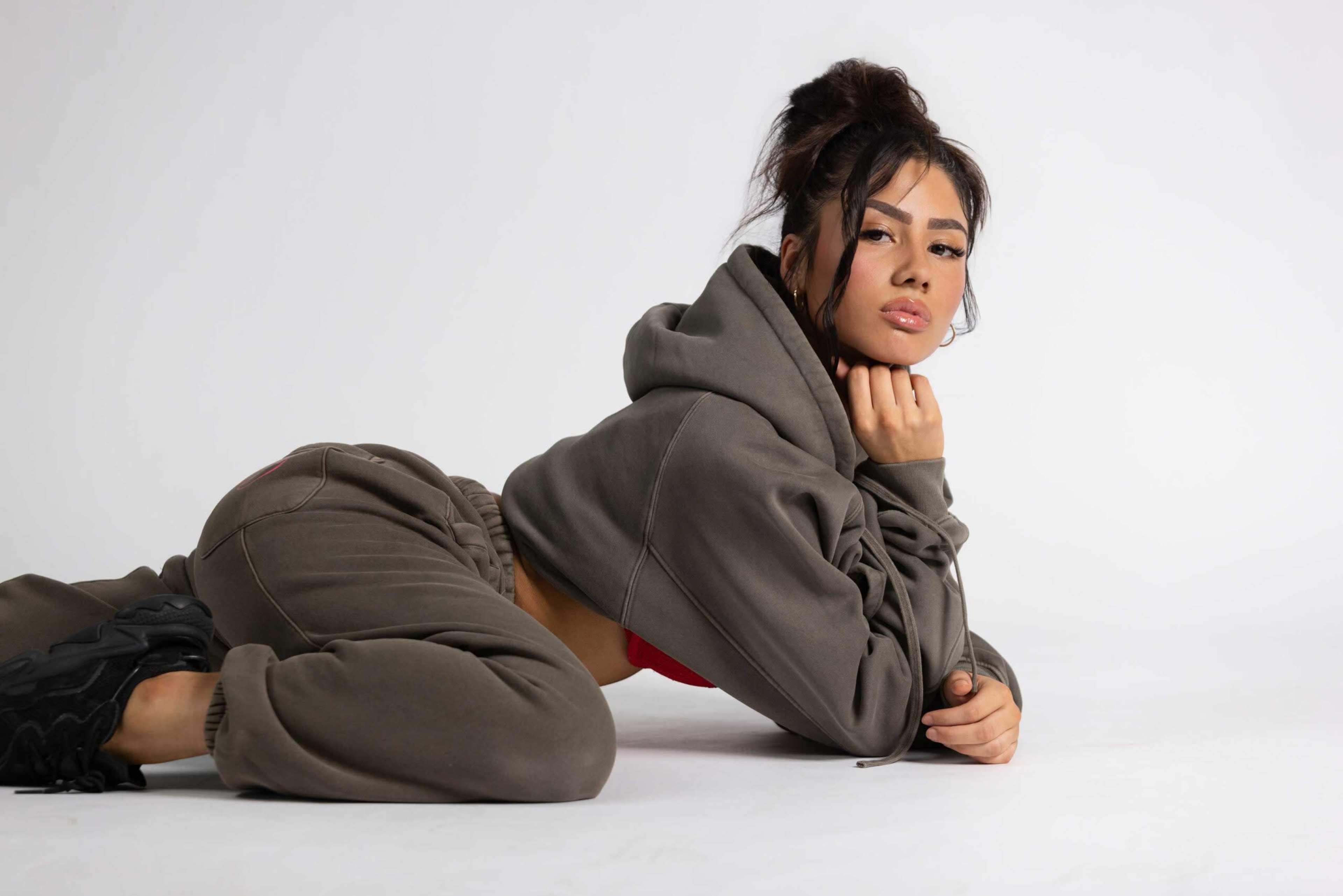 A person is posing on a white background, dressed in an olive green hoodie and matching sweatpants, with one hand resting on their chin.