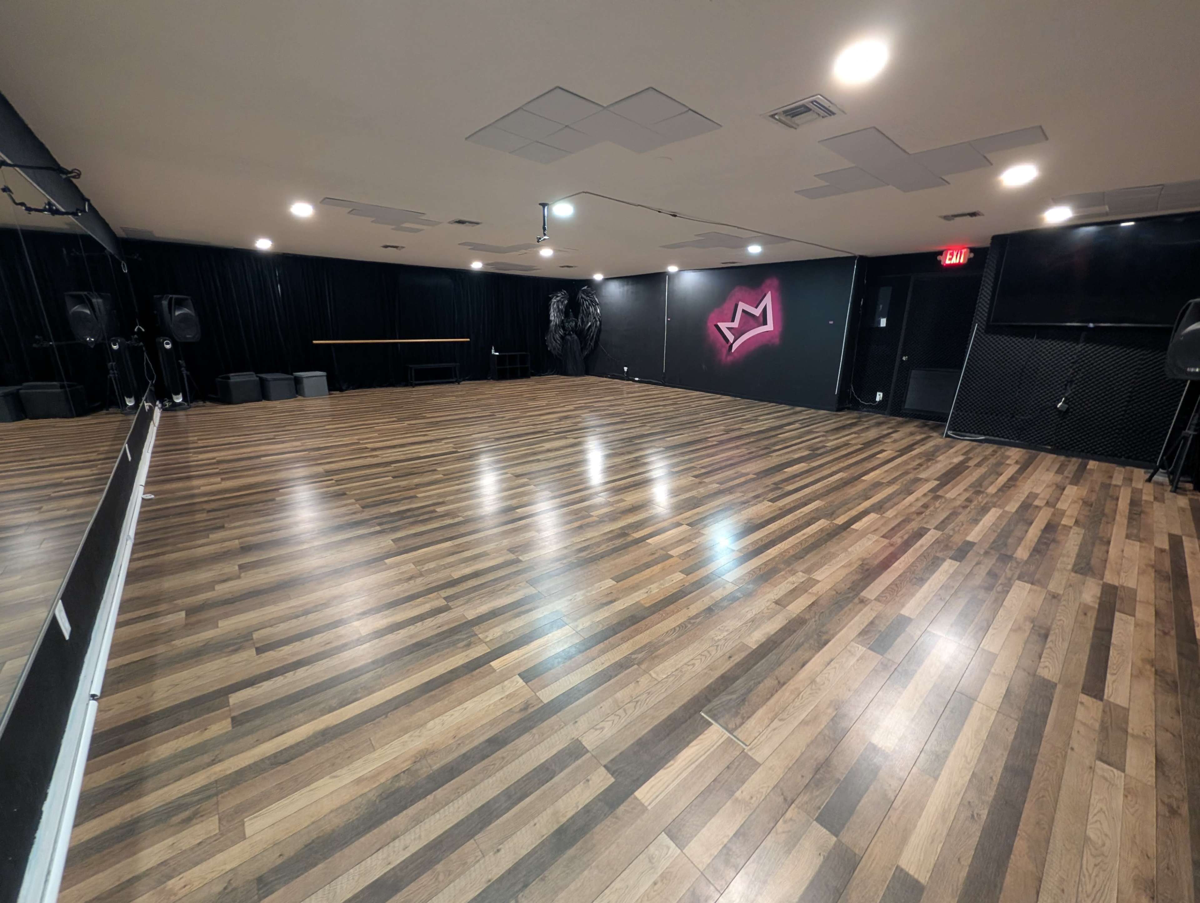 The image shows a spacious dance studio with wooden flooring, a large mirror on one wall, and a black backdrop featuring a stylized crown design.
