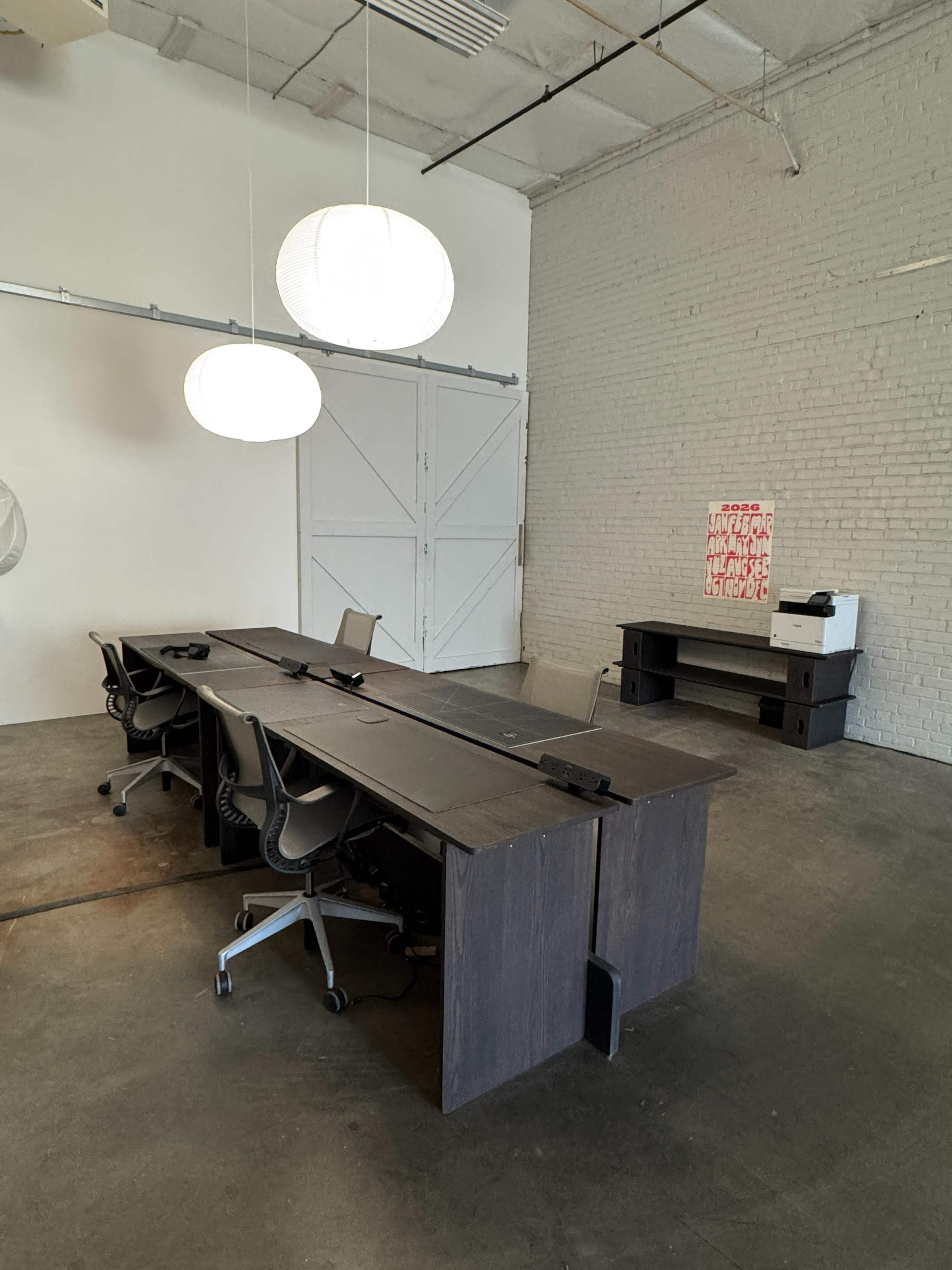 The image shows a minimalist office space with a long wooden table, several office chairs, and two pendant lights hanging from the ceiling.
