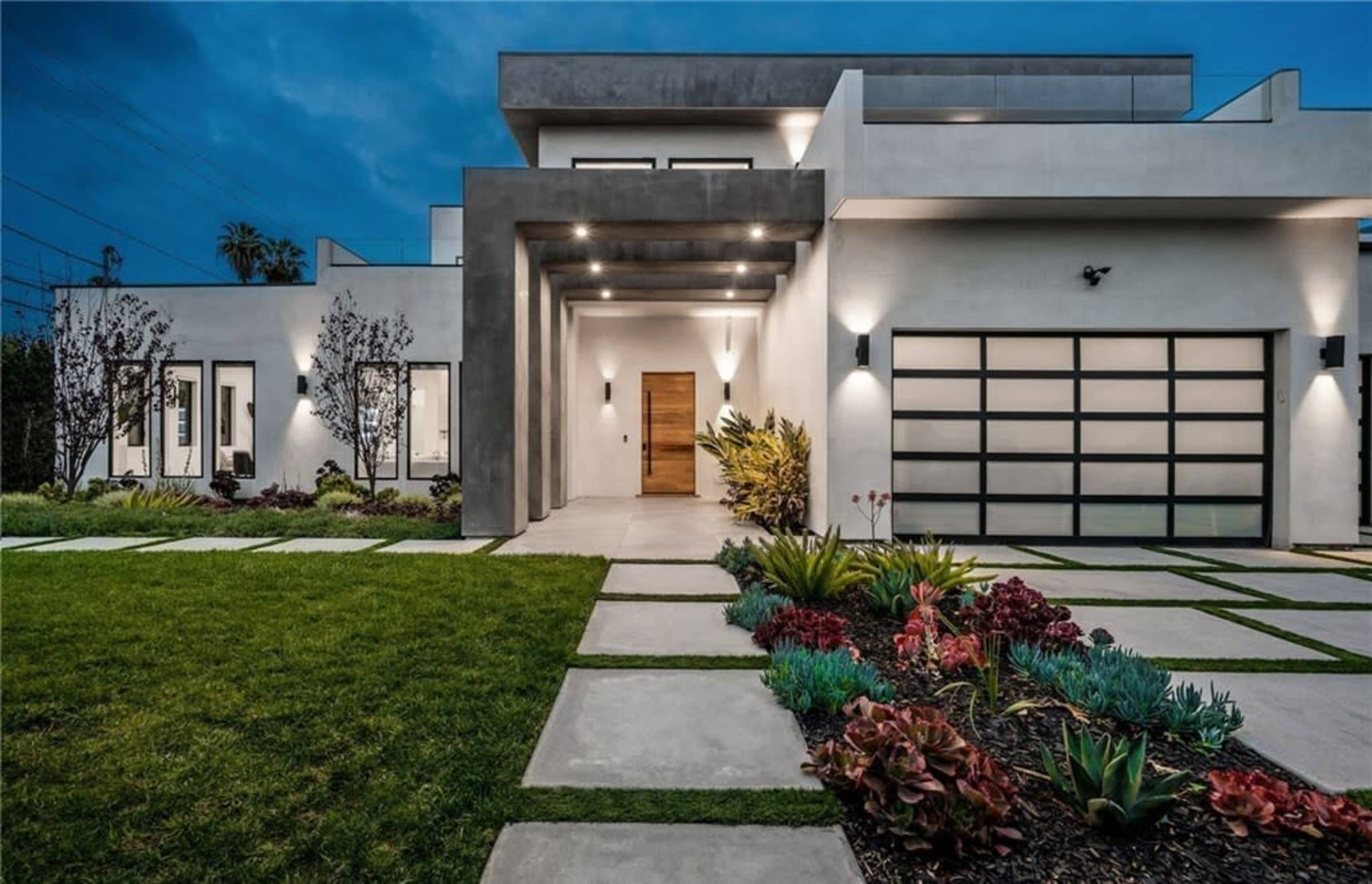 A modern house features a symmetrical entrance with a wooden door, large garage door, and landscaped garden pathways.