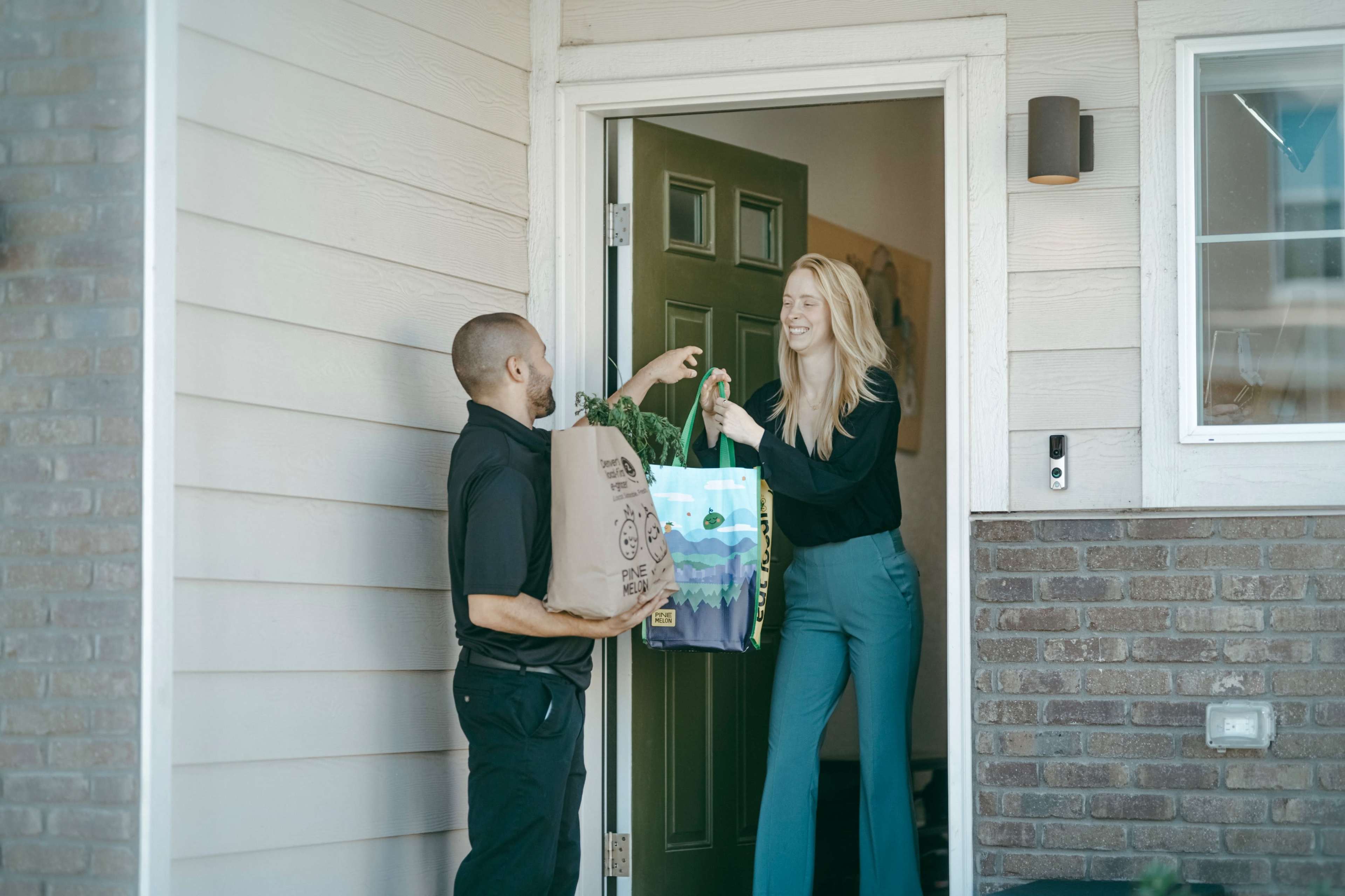 A delivery person hands bags of groceries to a woman standing in the doorway of a home.