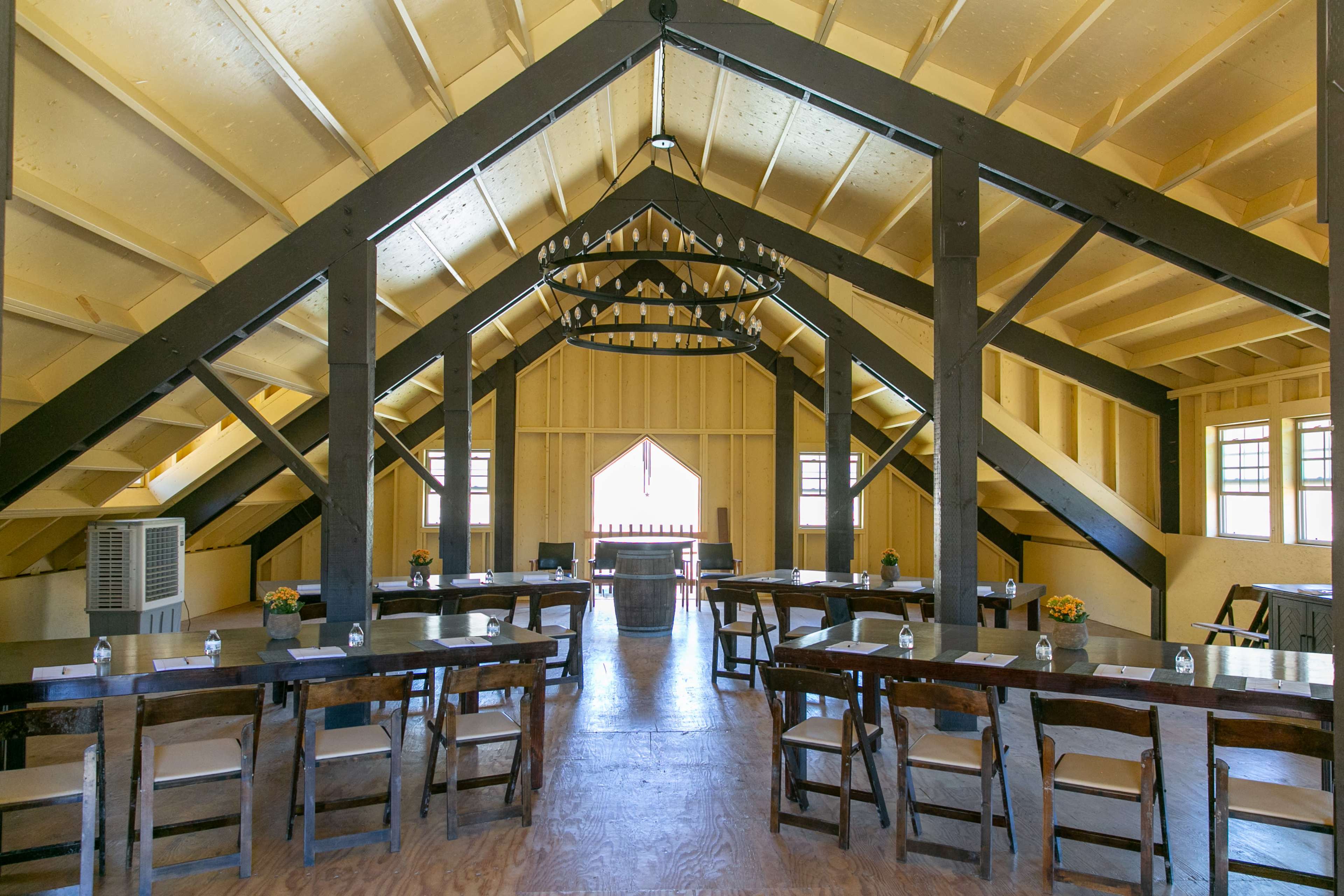 The image shows a spacious attic room with wooden beams, several tables arranged with floral centerpieces, and large windows allowing natural light.