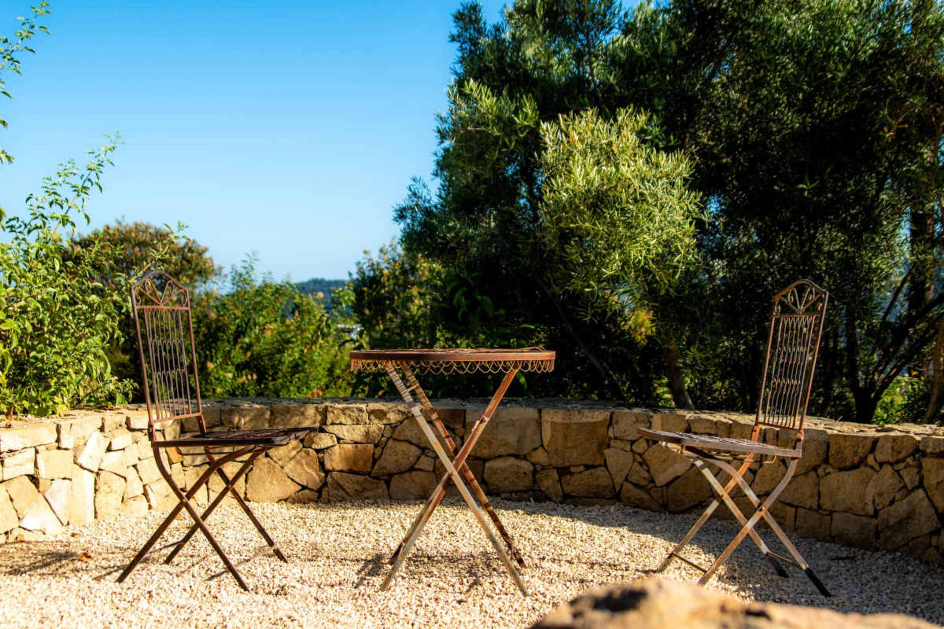 A small, circular table surrounded by two folding chairs is positioned on a gravel surface against a stone wall, with greenery in the background.