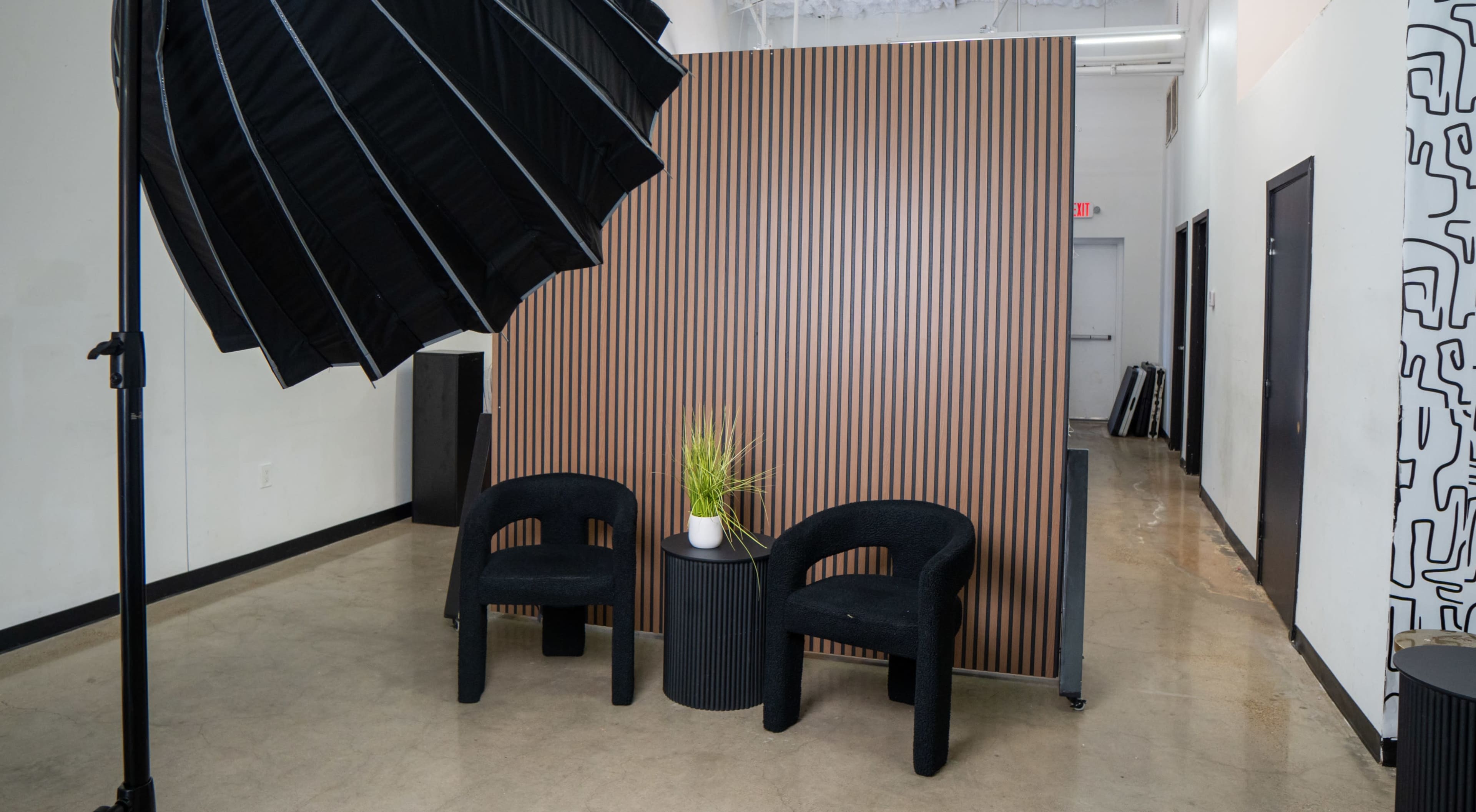 The image shows two black chairs in front of a wooden slatted wall, with a small plant on a round table, illuminated by a large softbox light.