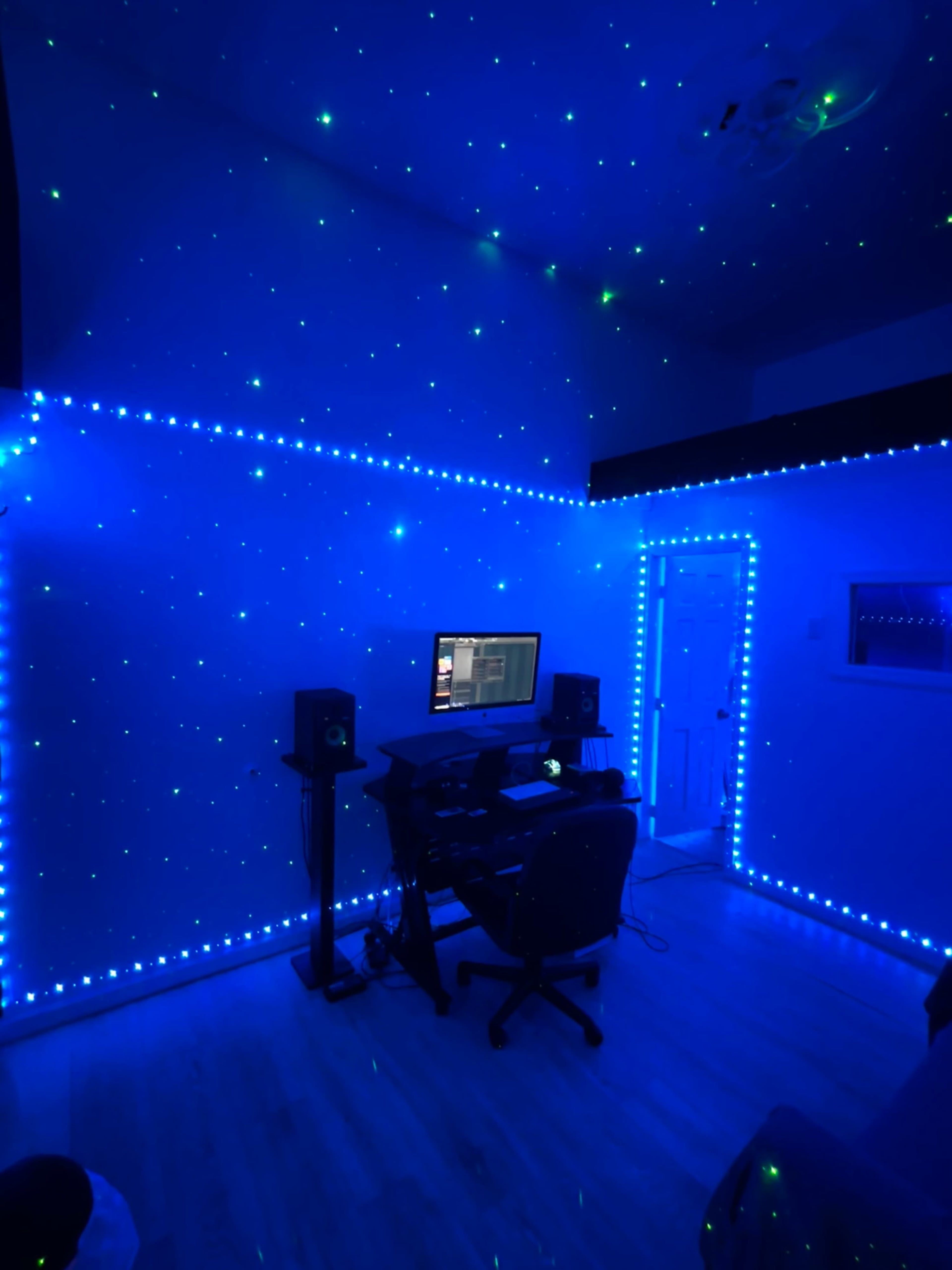 The image shows a dimly lit room with blue walls decorated with star-like lights, featuring a desk with a computer and audio speakers.