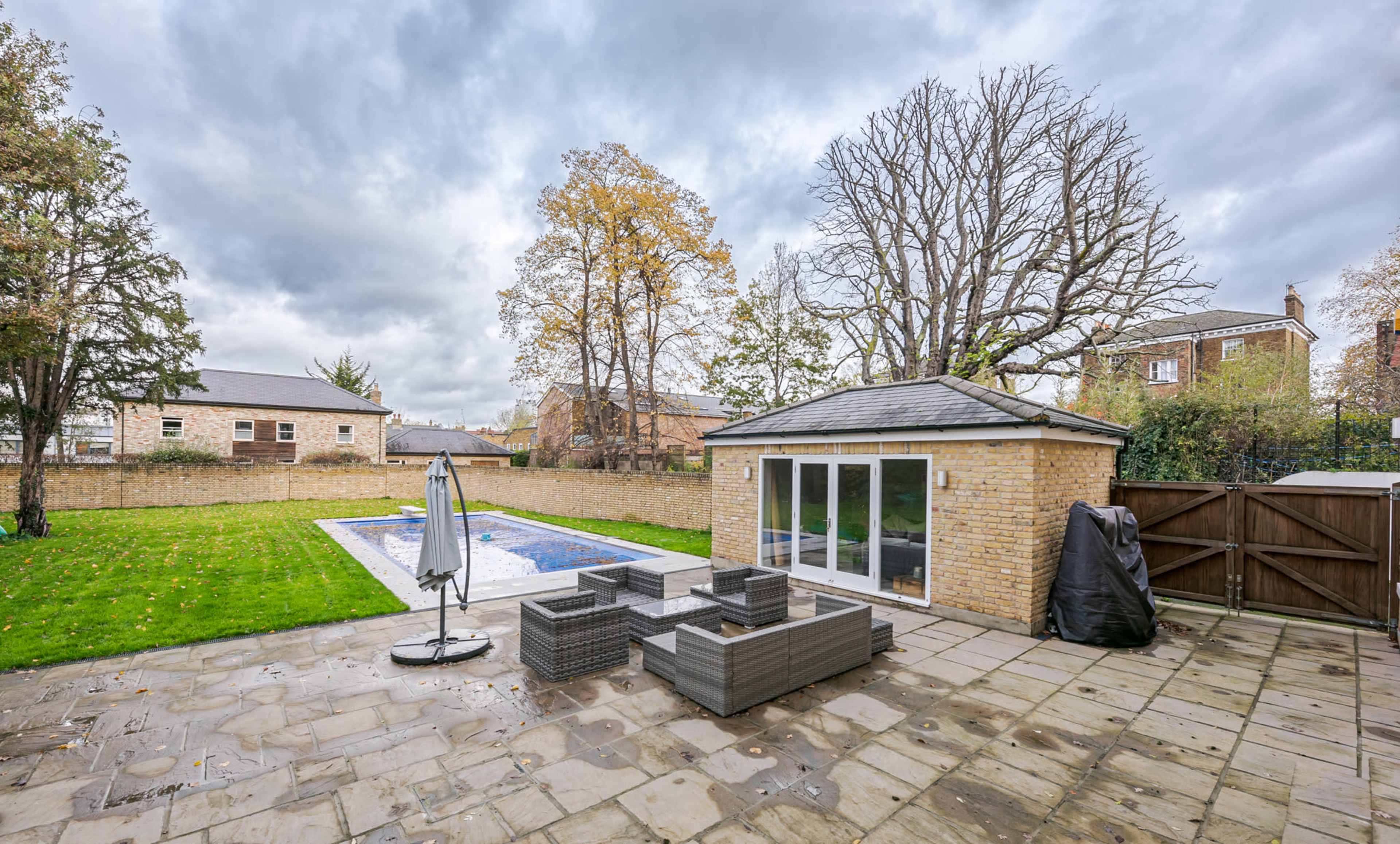 A backyard featuring a swimming pool, a patio area with outdoor furniture, and trees surrounded by a stone pathway.