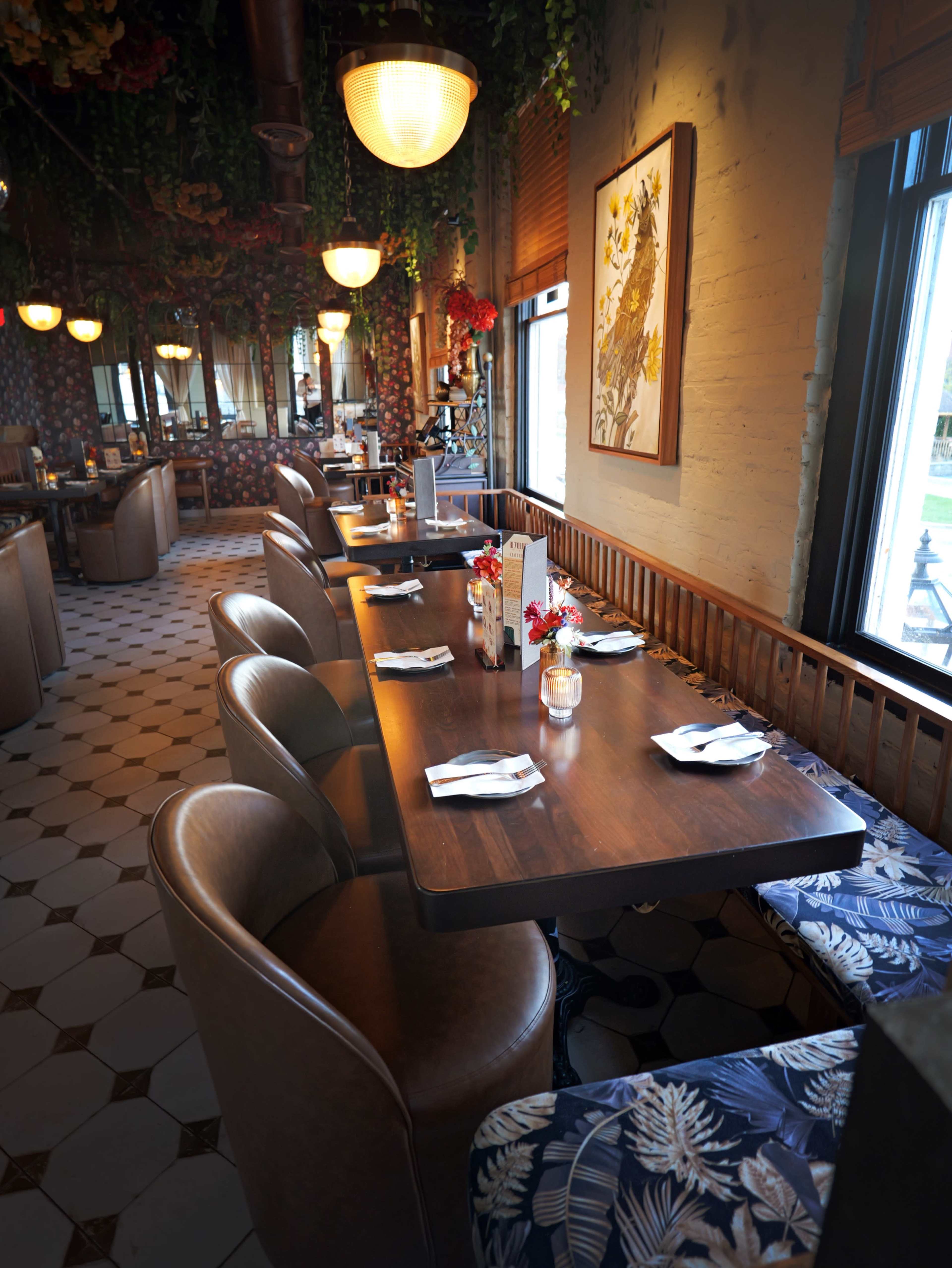 The image shows a well-decorated restaurant interior with several wooden tables set for dining and surrounded by floral arrangements and patterned seating.
