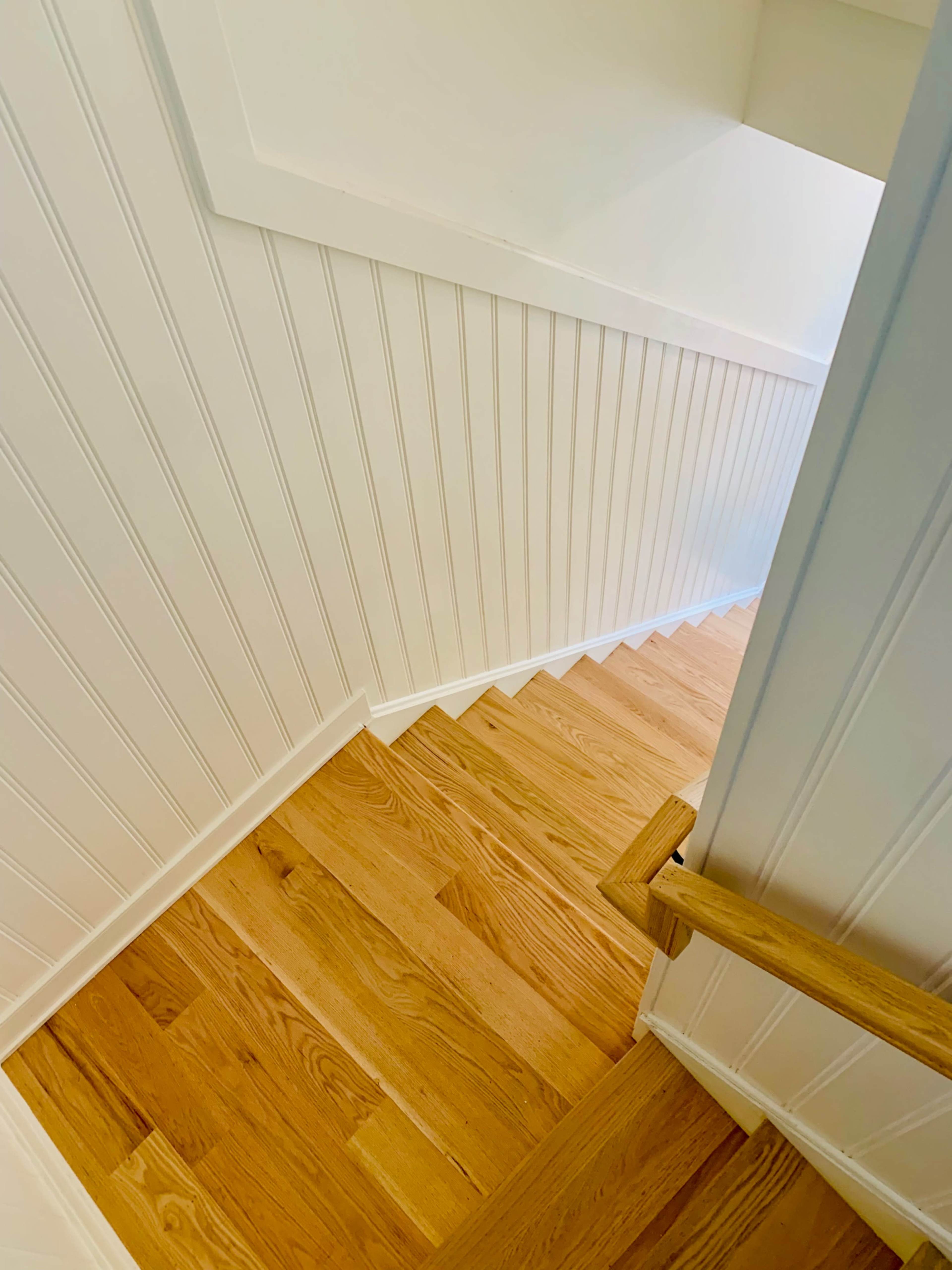 A view looking down a wooden staircase with a white paneled wall and light-colored flooring.