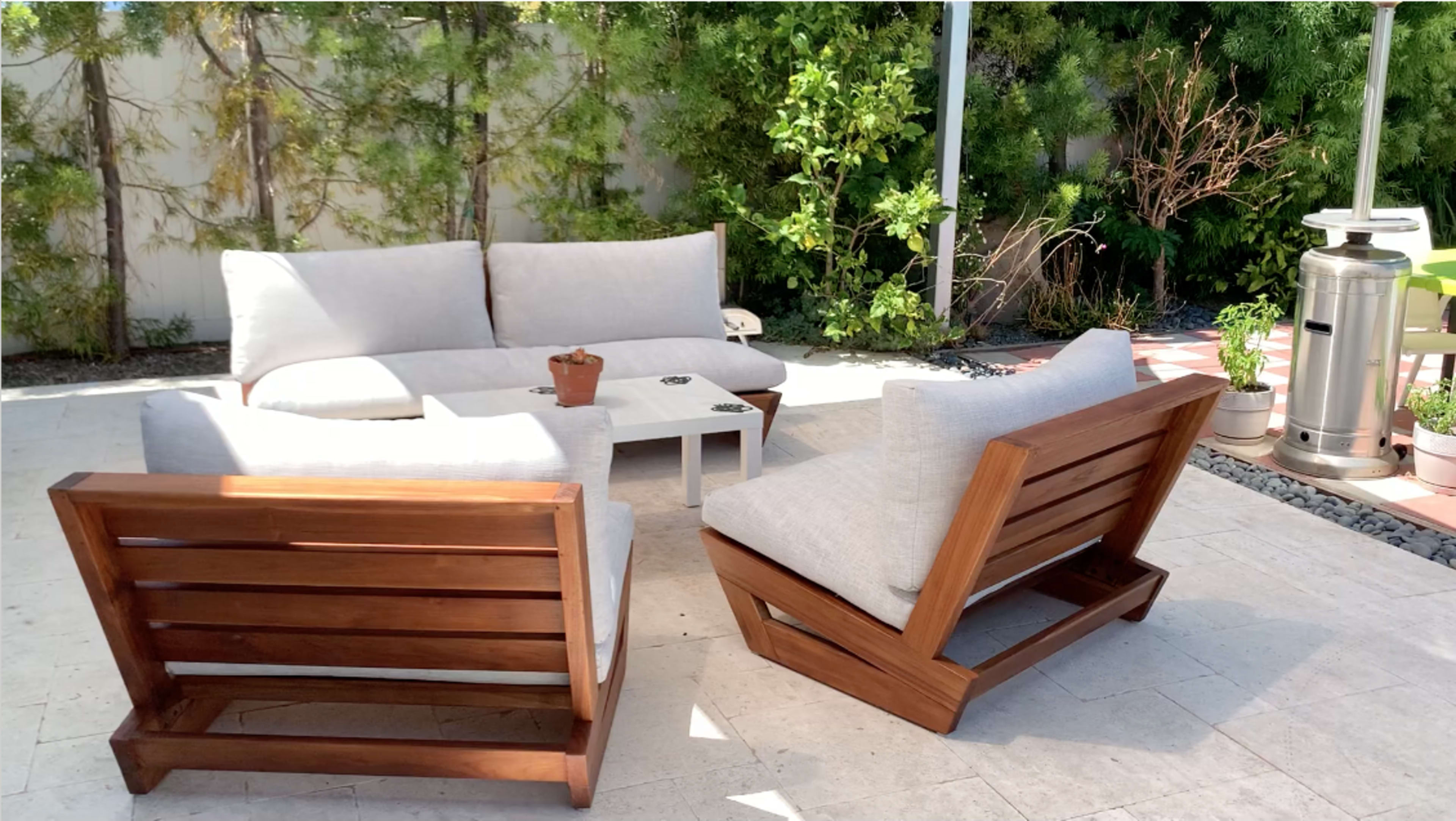 A set of modern outdoor furniture, including a sectional sofa and two armchairs, is arranged on a stone patio surrounded by greenery.