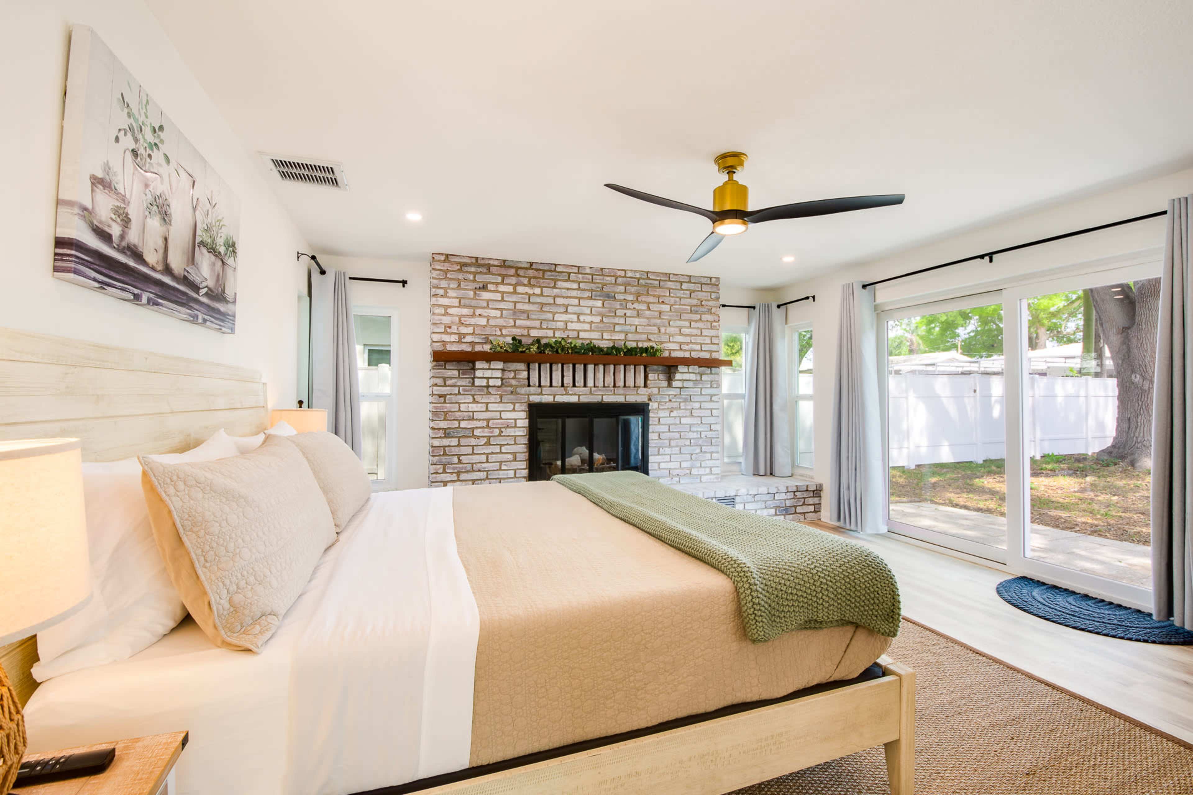A spacious bedroom features a large bed with neutral bedding, a brick fireplace, and sliding glass doors that open to a garden view.