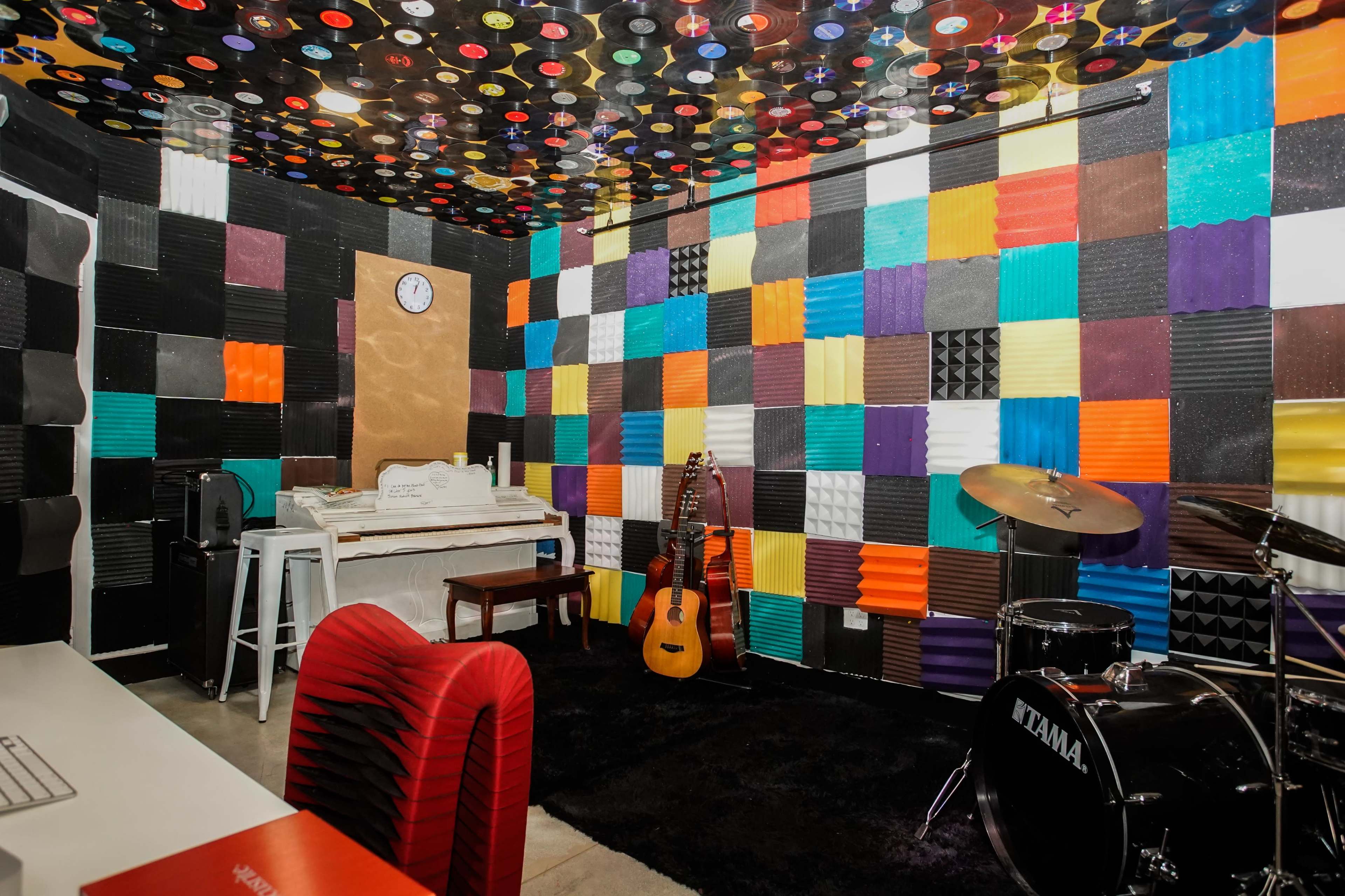 The image shows a colorful music studio with a wall covered in soundproofing panels, a keyboard, a guitar, a drum set, and a clock.
