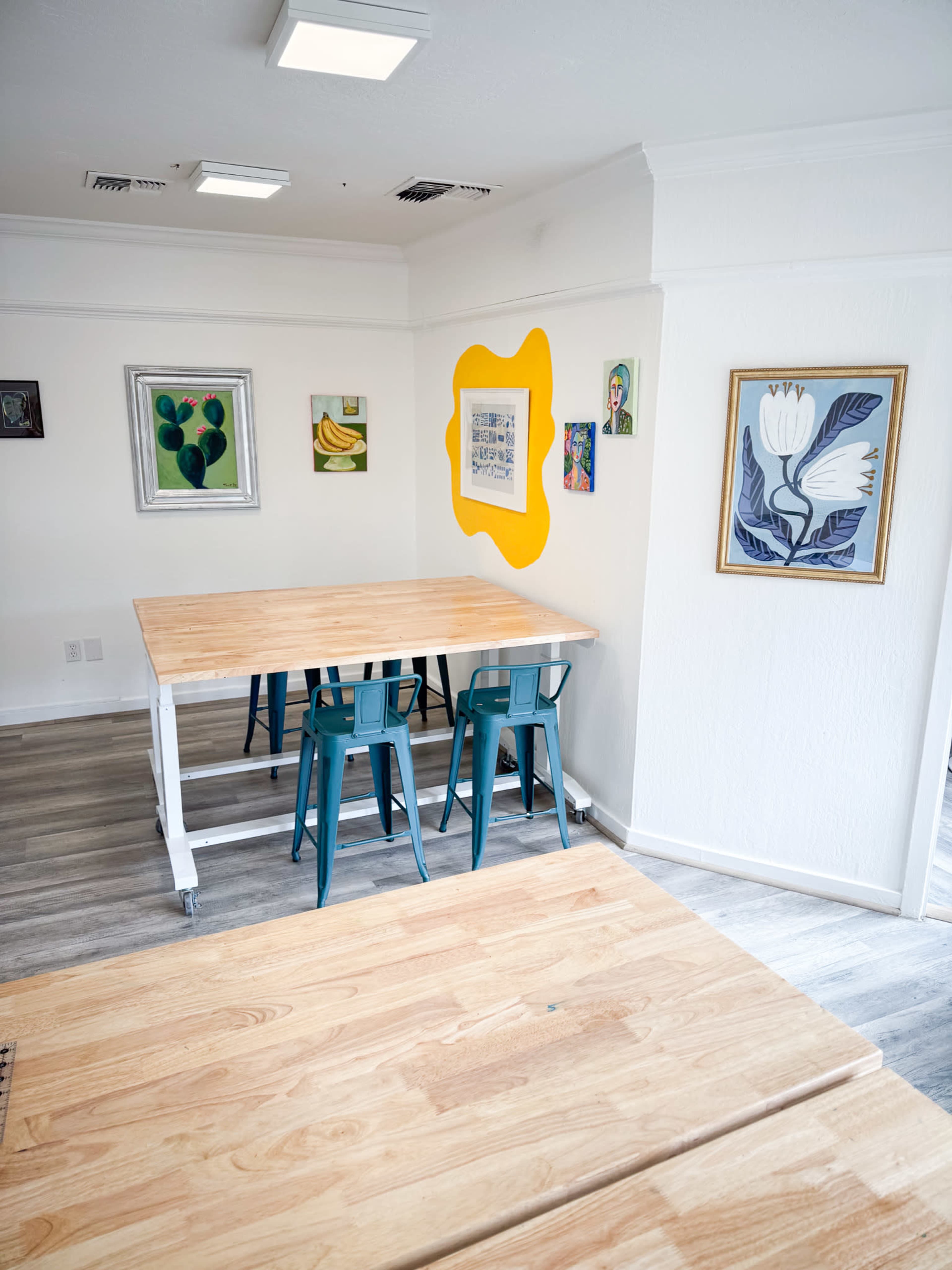A bright room featuring a wooden table and teal chairs, with colorful artworks hanging on the walls.